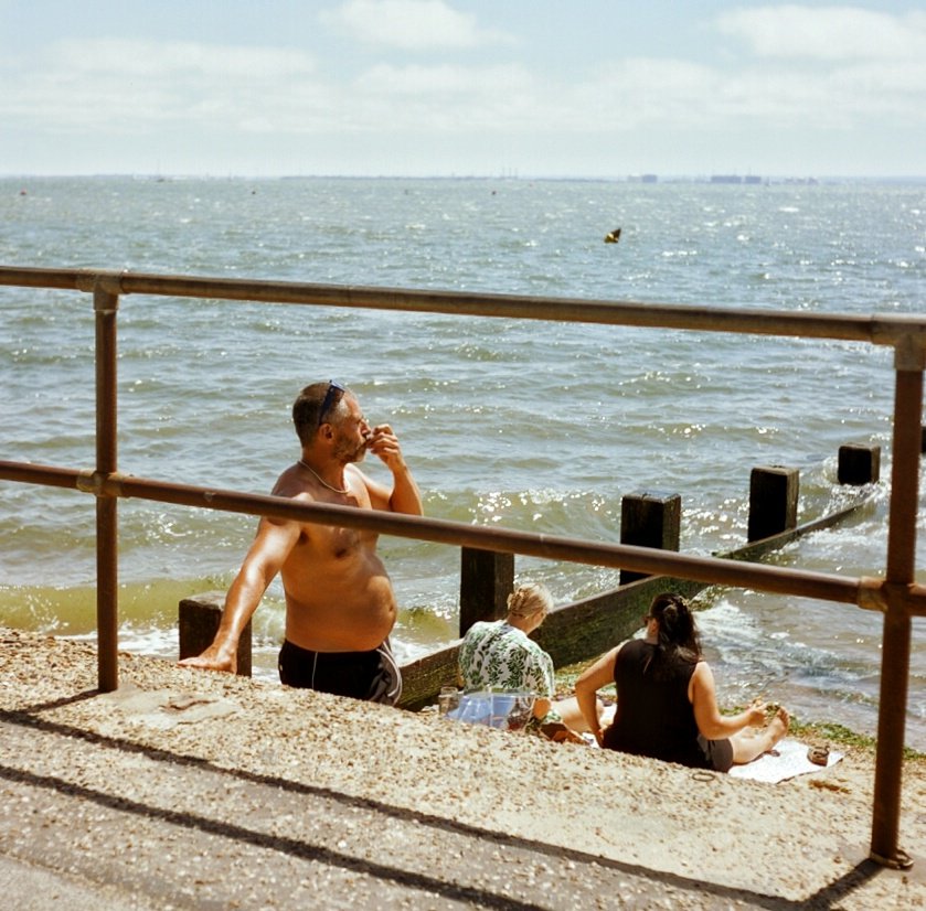 People relaxing by the water at a beach, with a man standing shirtless and two women sitting on a towel near the shoreline, while others are in the water and looking out at the sea.