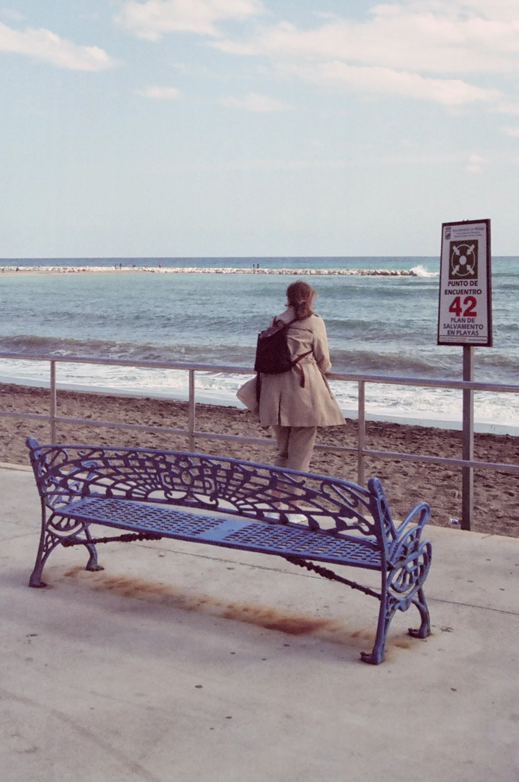 A woman in a beige coat and pants stands by a railing, looking at the ocean. There is a decorative blue bench in the foreground, and the beach with a sign is visible in the background.