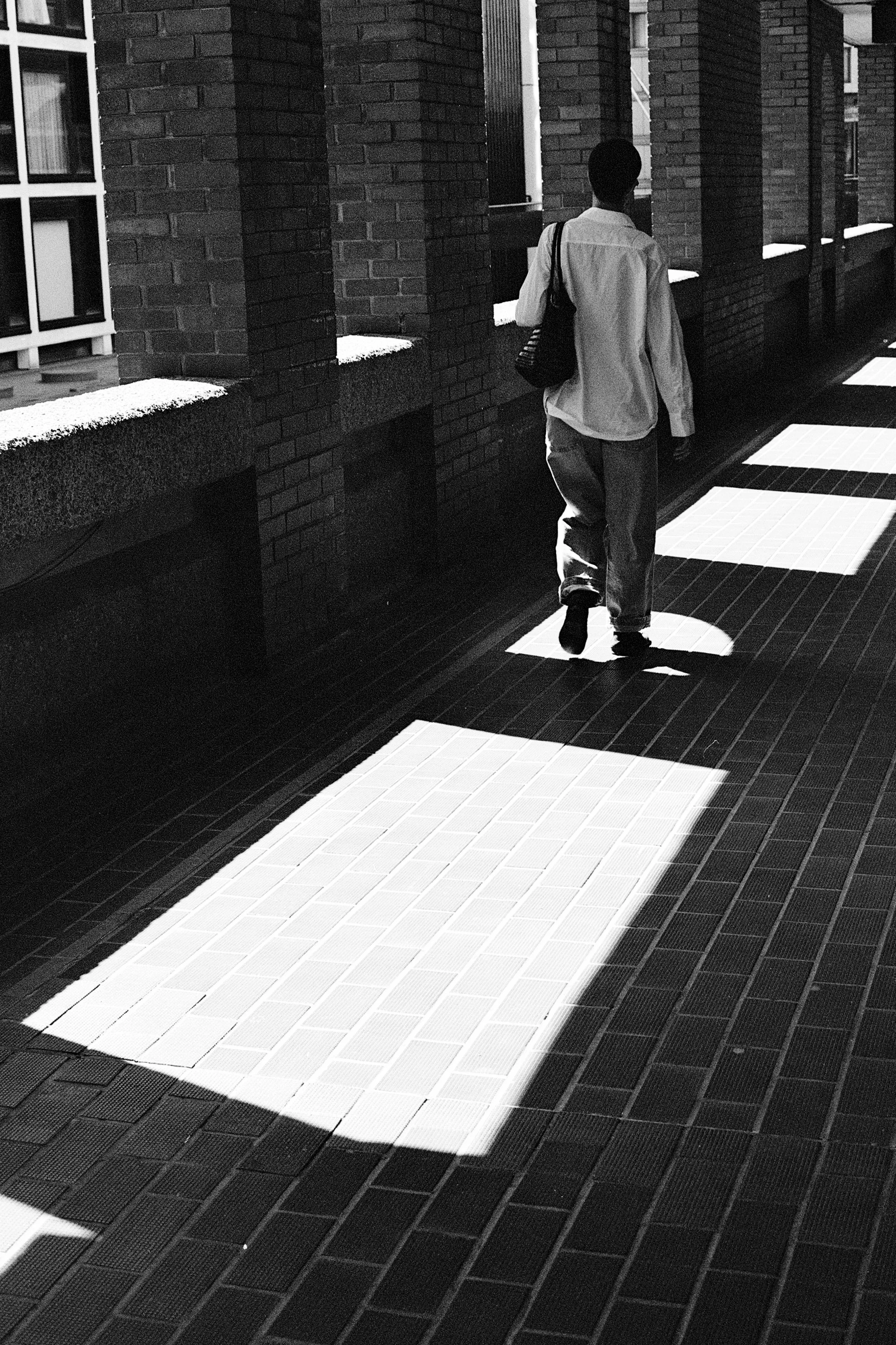 A person walking along a brick sidewalk with sunlight creating rectangular patches of shadow and light.