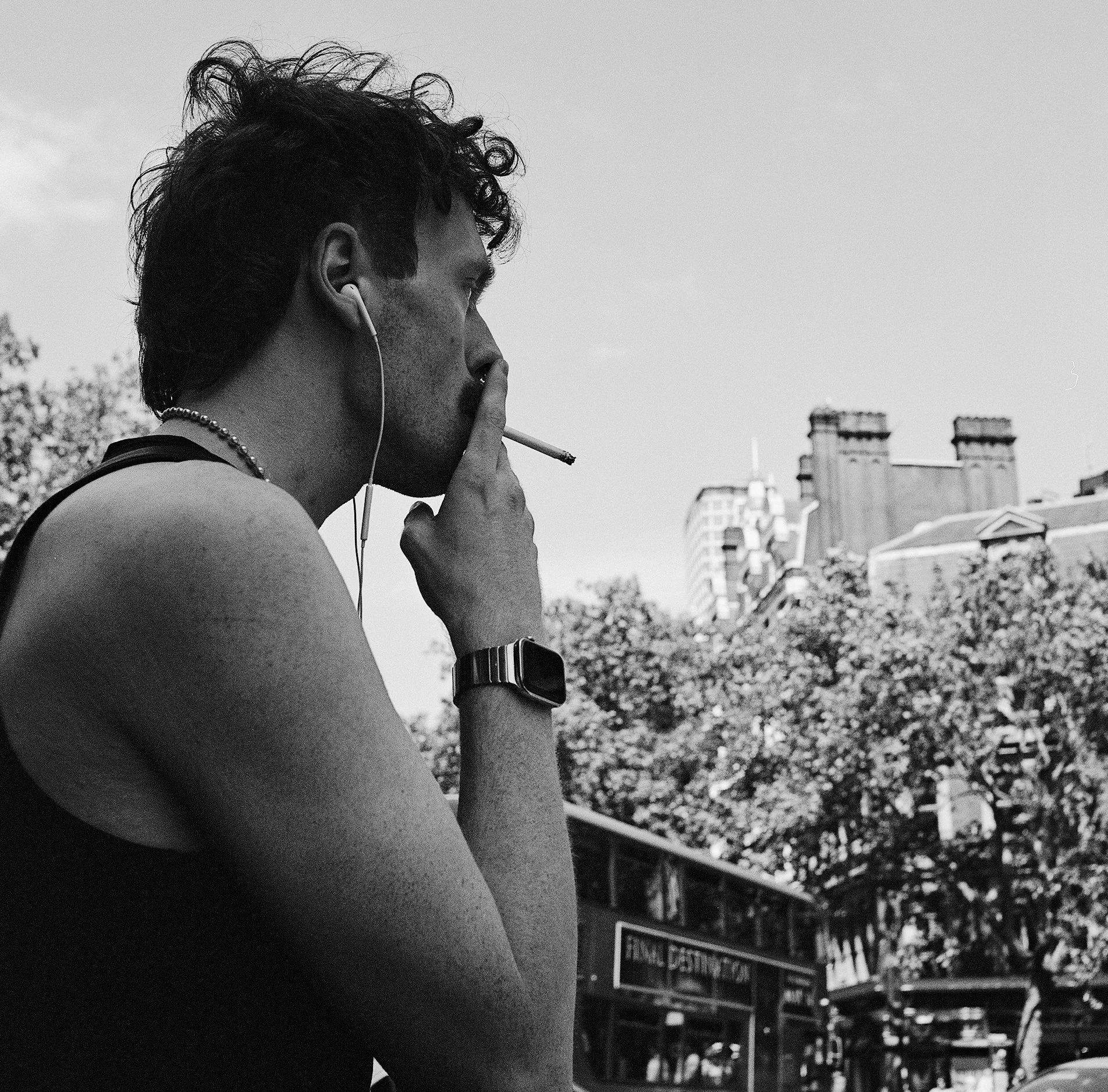 A black and white photo of a man with curly hair smoking a cigarette outdoors in a city, wearing earphones, a necklace, a wristwatch, and a sleeveless top, with trees and city buildings in the background.
