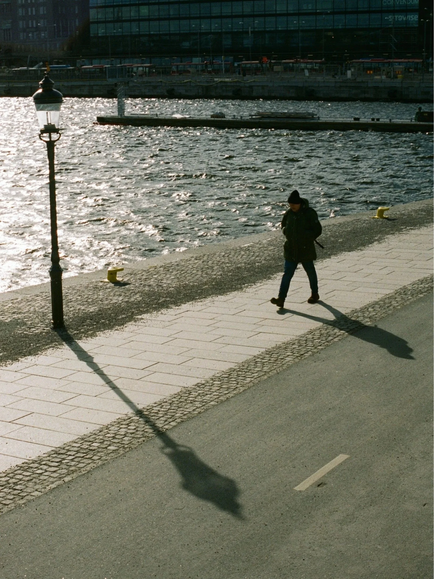 A person in a dark coat and beanie walking along a riverside promenade, with a lamppost casting a long shadow on the pavement. The river has small waves, and there are boats and buildings visible in the background under sunlight.