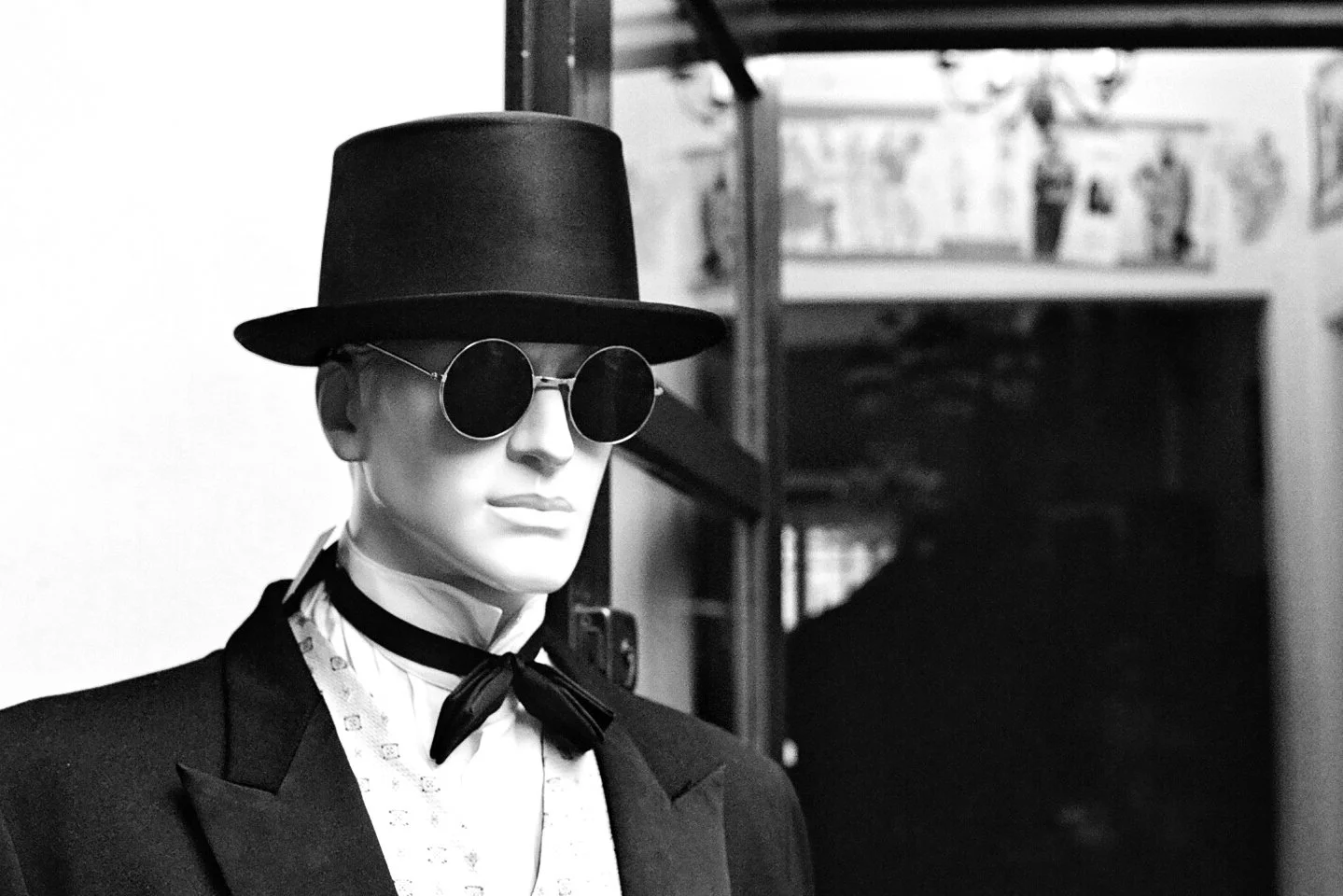 Black and white photo of a mannequin wearing a round sunglasses, a black top hat, a bow tie, and a formal jacket.