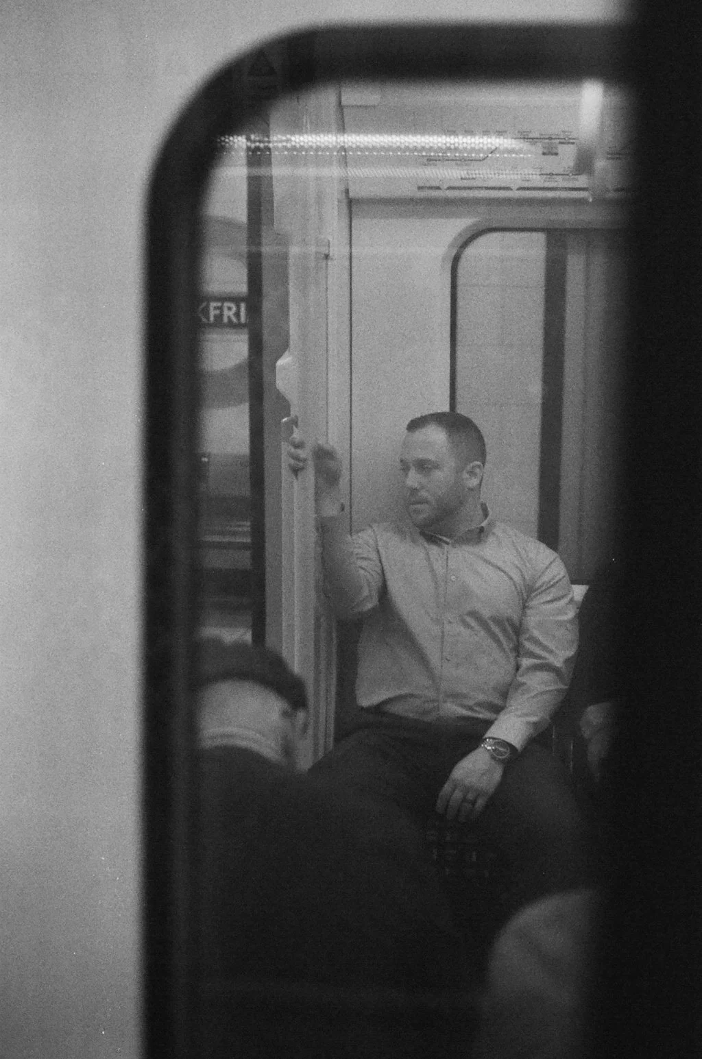 A man with short hair, wearing a button-up shirt and a wristwatch, sitting and looking to his left during a train or subway ride, seen through a window or mirror.