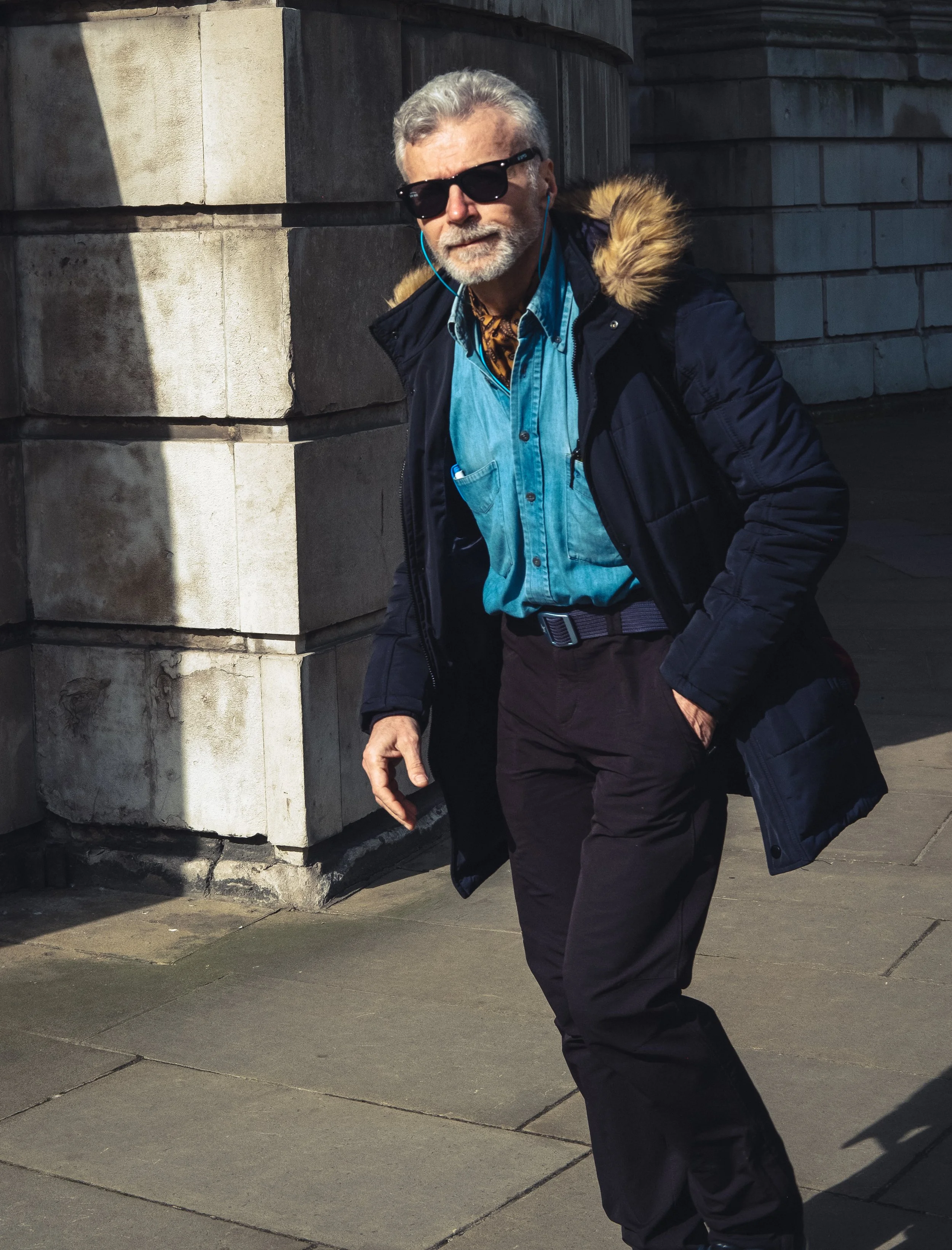 An older man with gray hair and beard wearing black sunglasses, a blue denim shirt, dark pants, and a black coat with a fur-lined hood, walking on a city sidewalk near a stone building.