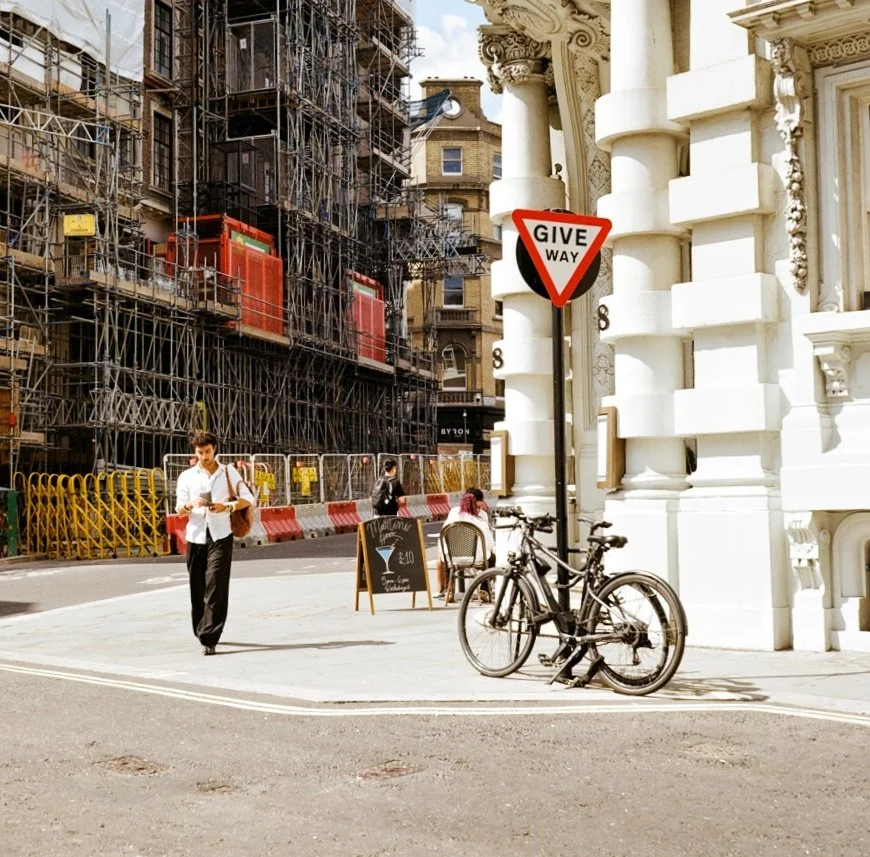 Street scene featuring a man walking with a bag, two bicycles parked against a building, a blackboard menu, a stop sign, and construction scaffolding on nearby buildings.