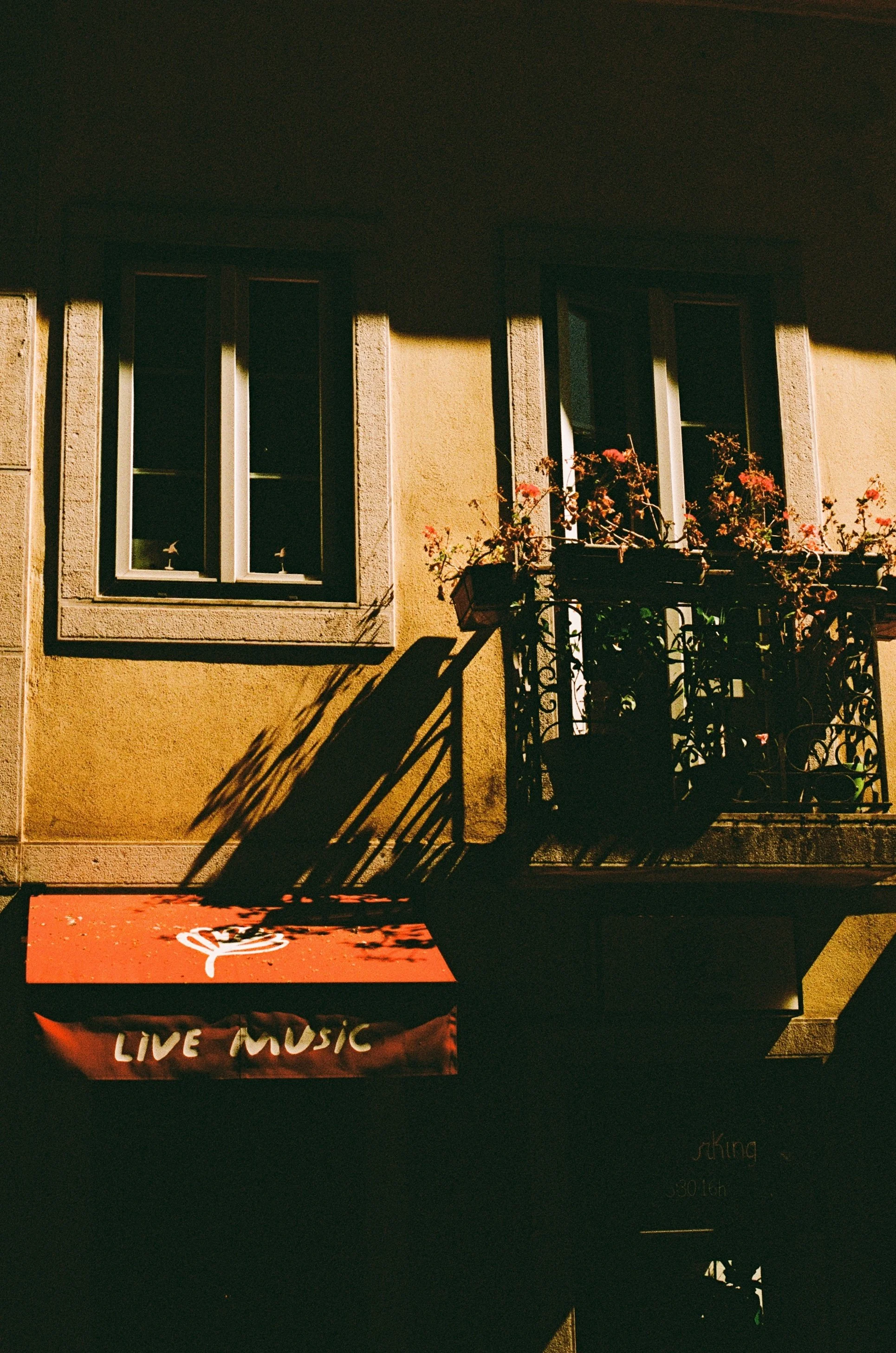 Sunlit building with two windows, potted plants on a balcony, and a red awning with 'LIVE MUSIC' written on it.