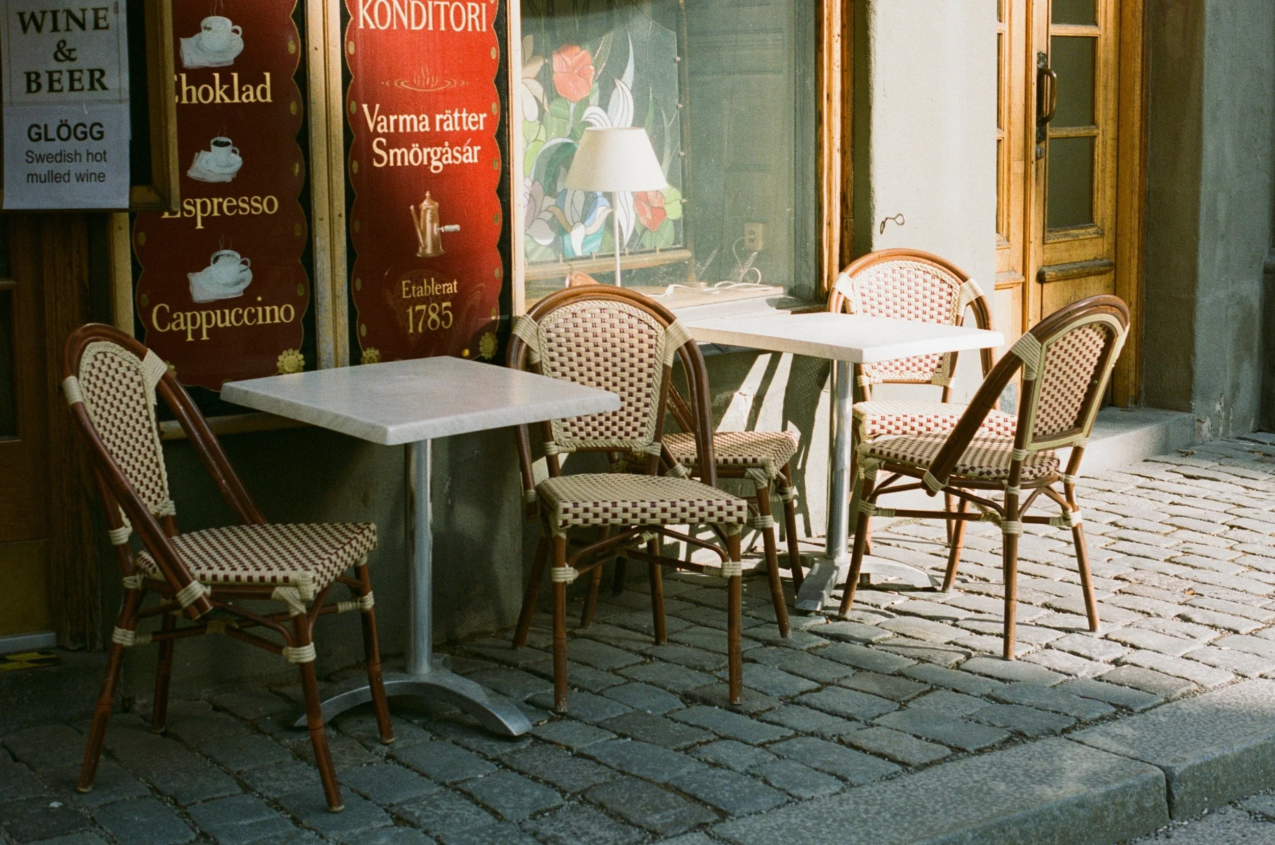 Outdoor seating with four chairs around two white tables outside a cafe, with signs advertising wine, beer, coffee, and baked goods in Swedish.