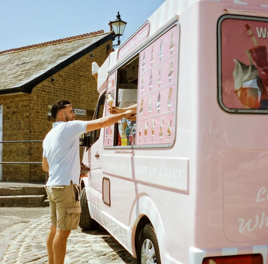 A man in a white shirt and beige shorts getting ice cream from a pink ice cream truck with a service window, on a cobblestone street in front of a brick building with a sign for toilets.
