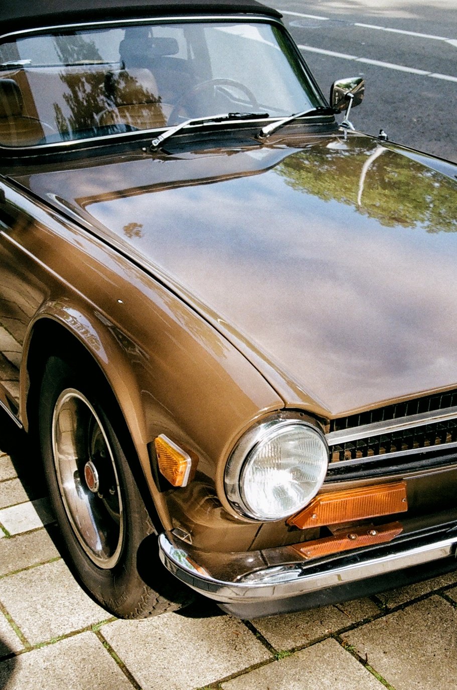 A vintage brown car parked on a brick sidewalk, showing the front left side with the headlight, wheel, and a reflection of trees and the sky on the windshield and hood.