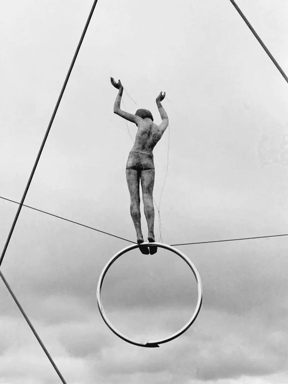 A black and white photograph of a statue of a woman with arms raised, standing on a large hoop with a cloudy sky in the background.