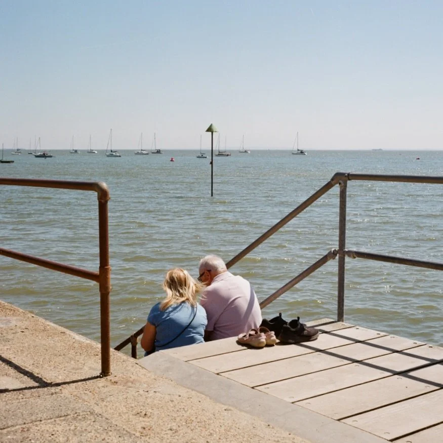 An older man and a young girl sitting on a pier by the water, with shoes beside them and sailboats in the distance.