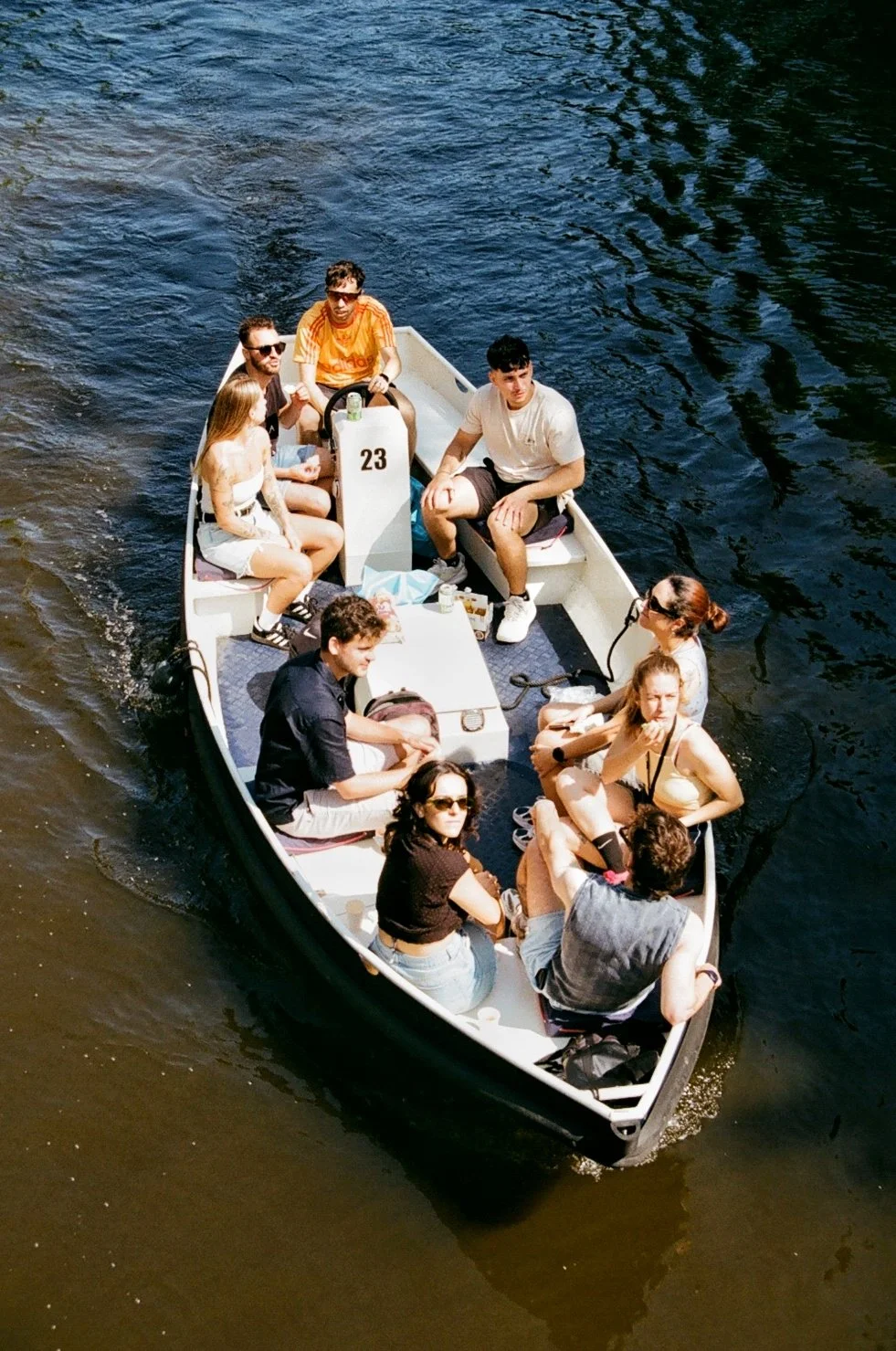 A group of people riding a small boat on a dark river, some are seated and chatting, enjoying the sunny day.