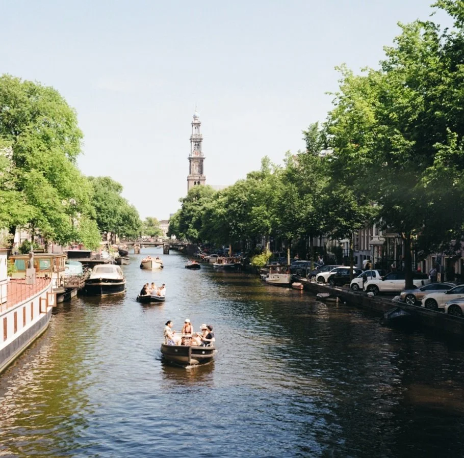 Boats floating on a canal lined with green trees and parked cars, with a tall historic tower in the background on a sunny day.