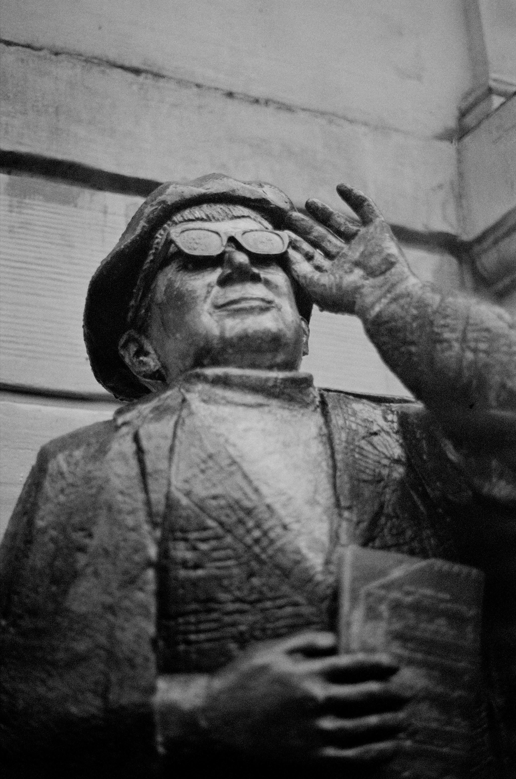 A black and white photograph of a statue of a person wearing glasses and a hat, with one hand raised to their glasses and holding a book or paper in the other hand.