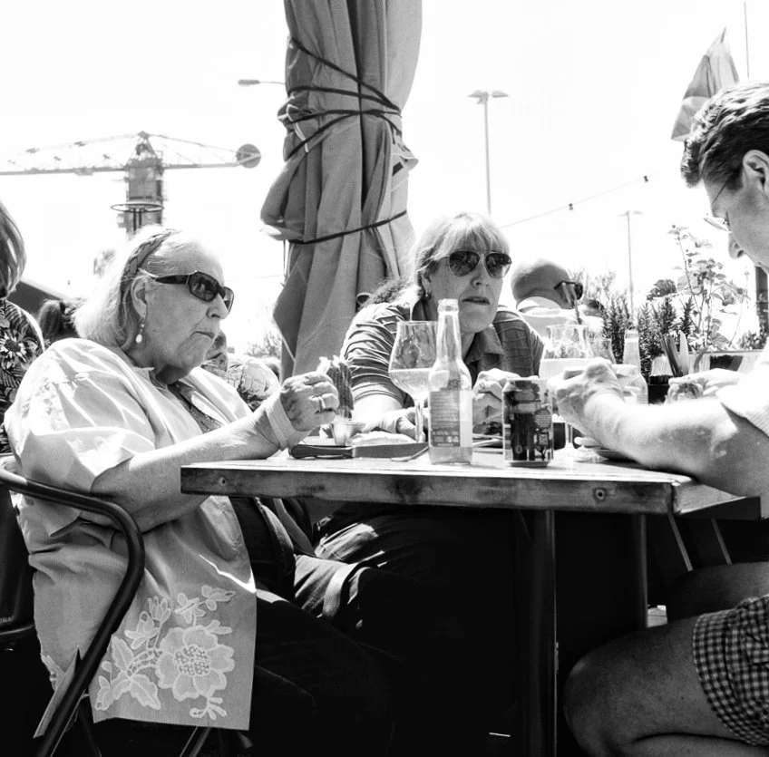 Four women and one man sitting at an outdoor table with drinks, engaged in conversation, in a sunny setting with a large umbrella and construction crane in the background.