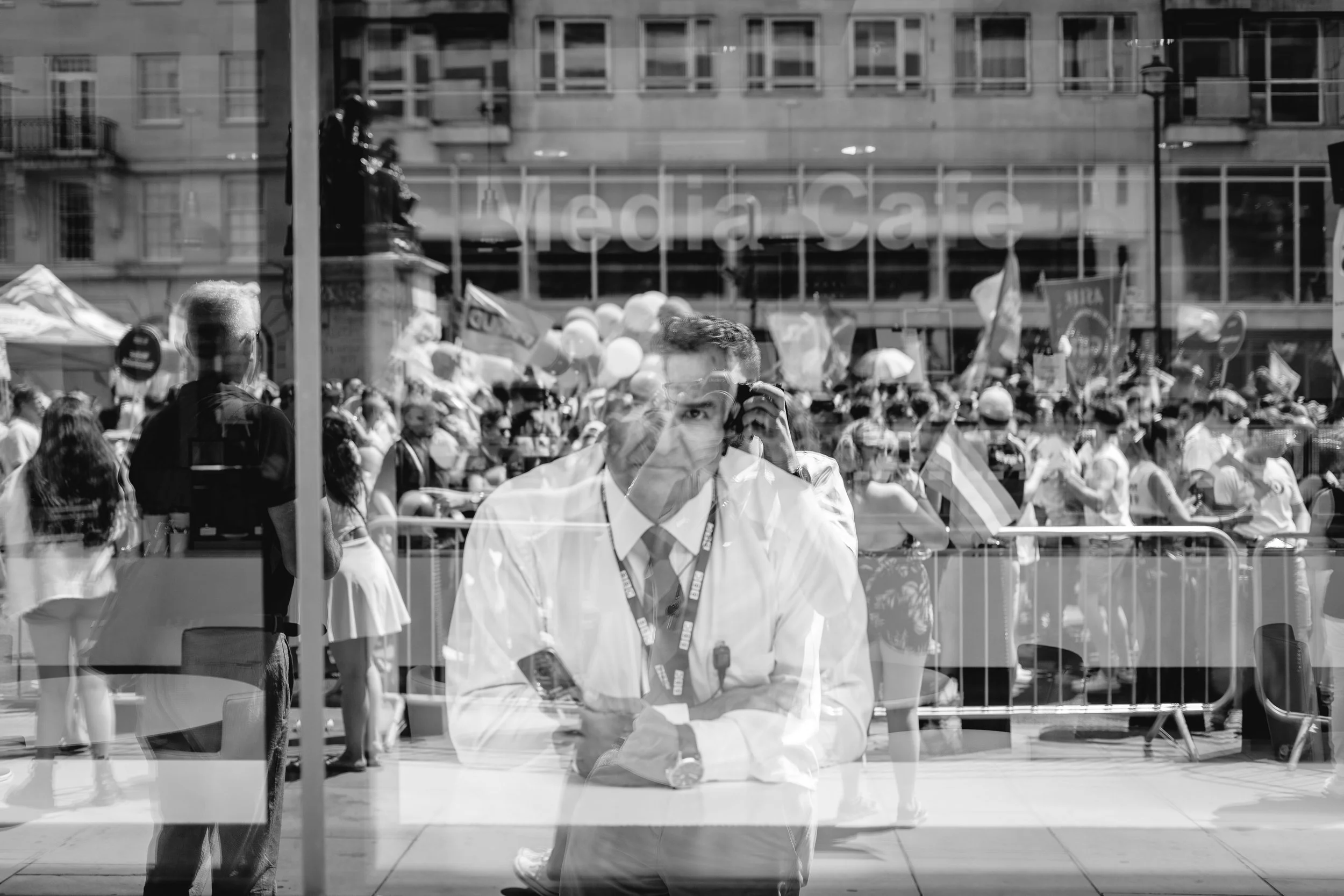 A reflection of a man in a white shirt and tie with an event or protest crowd behind him, seen through a glass window.