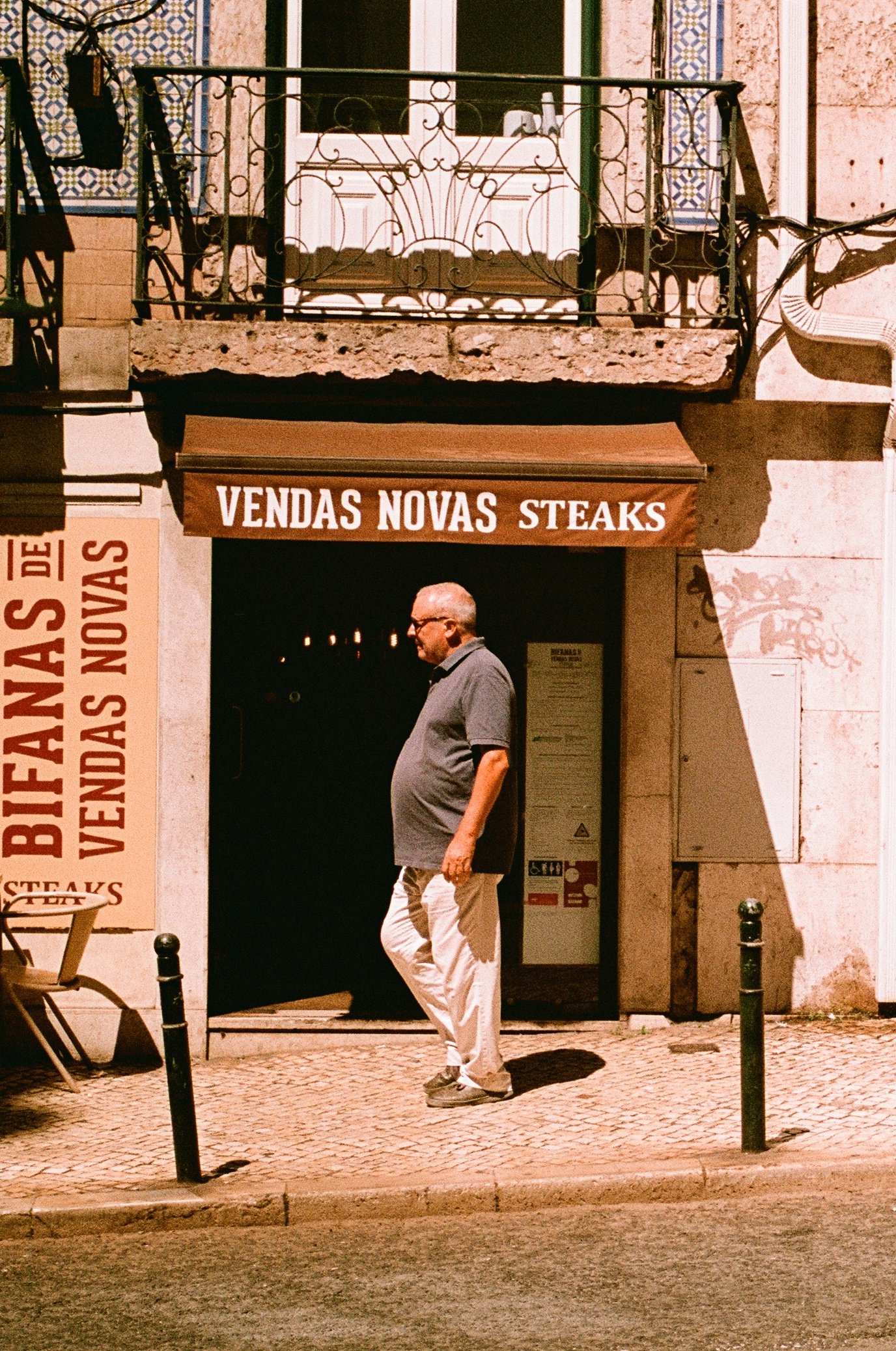 A man walking past a restaurant with signs advertising 'Vendas Novas Steaks' and 'Bifanas de Vendas Novas' on a cobblestone sidewalk.