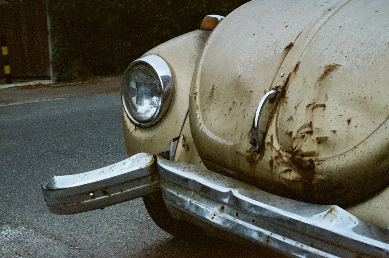 Close-up of a vintage beige car with a rusted front bumper and dirt on the hood, parked on a street.