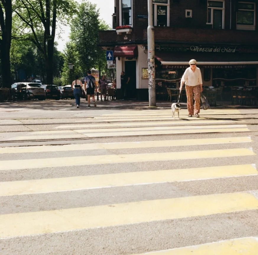 An elderly woman walking her small dog across a crosswalk on a city street, with a restaurant and parked cars in the background.