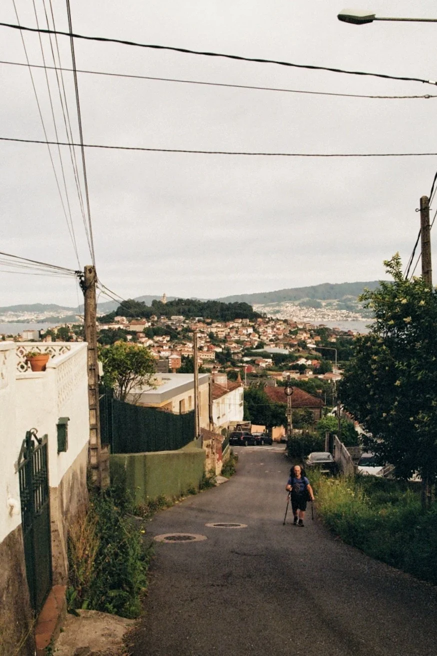 A woman walking up a sloped residential street with houses and trees, overlooking a hilly cityscape with numerous buildings and a distant hill with a monument or statue on top, under a cloudy sky.