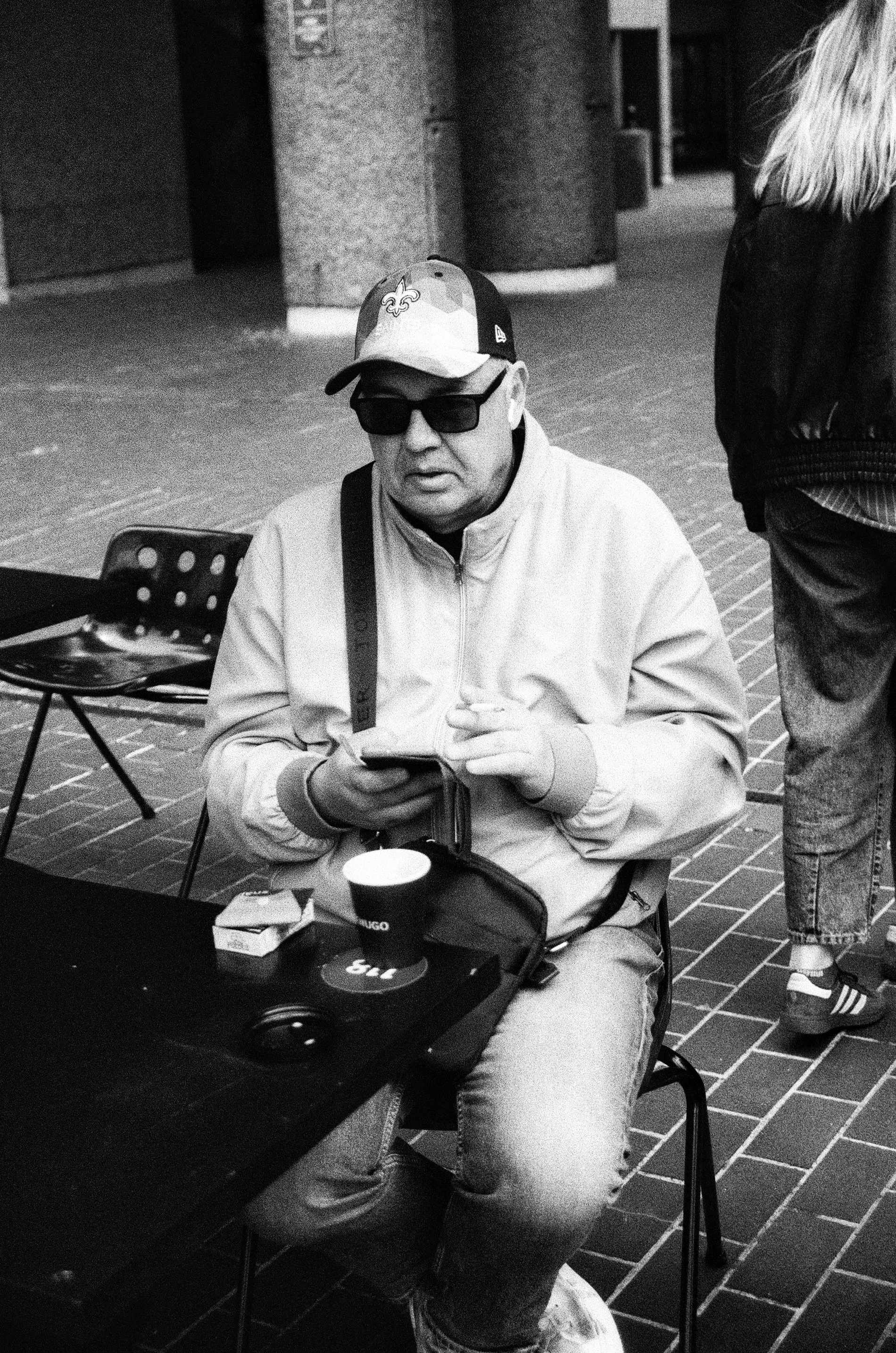 A man in a baseball cap, sunglasses, and a light jacket sitting at a table outdoors, looking at his phone. There is a paper cup, a small box, and a pair of headphones on the table.