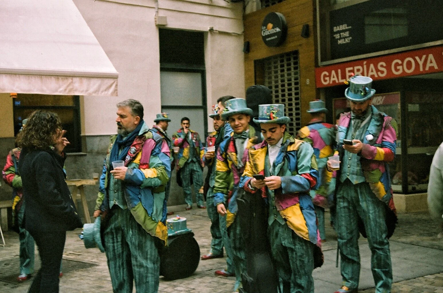 A group of people dressed in colorful, patchwork costumes and top hats, some using their phones, gathered outside a building with a sign reading 'Galerías Goya' and a Guiness advertisement, while a woman talks to a man holding a drink.