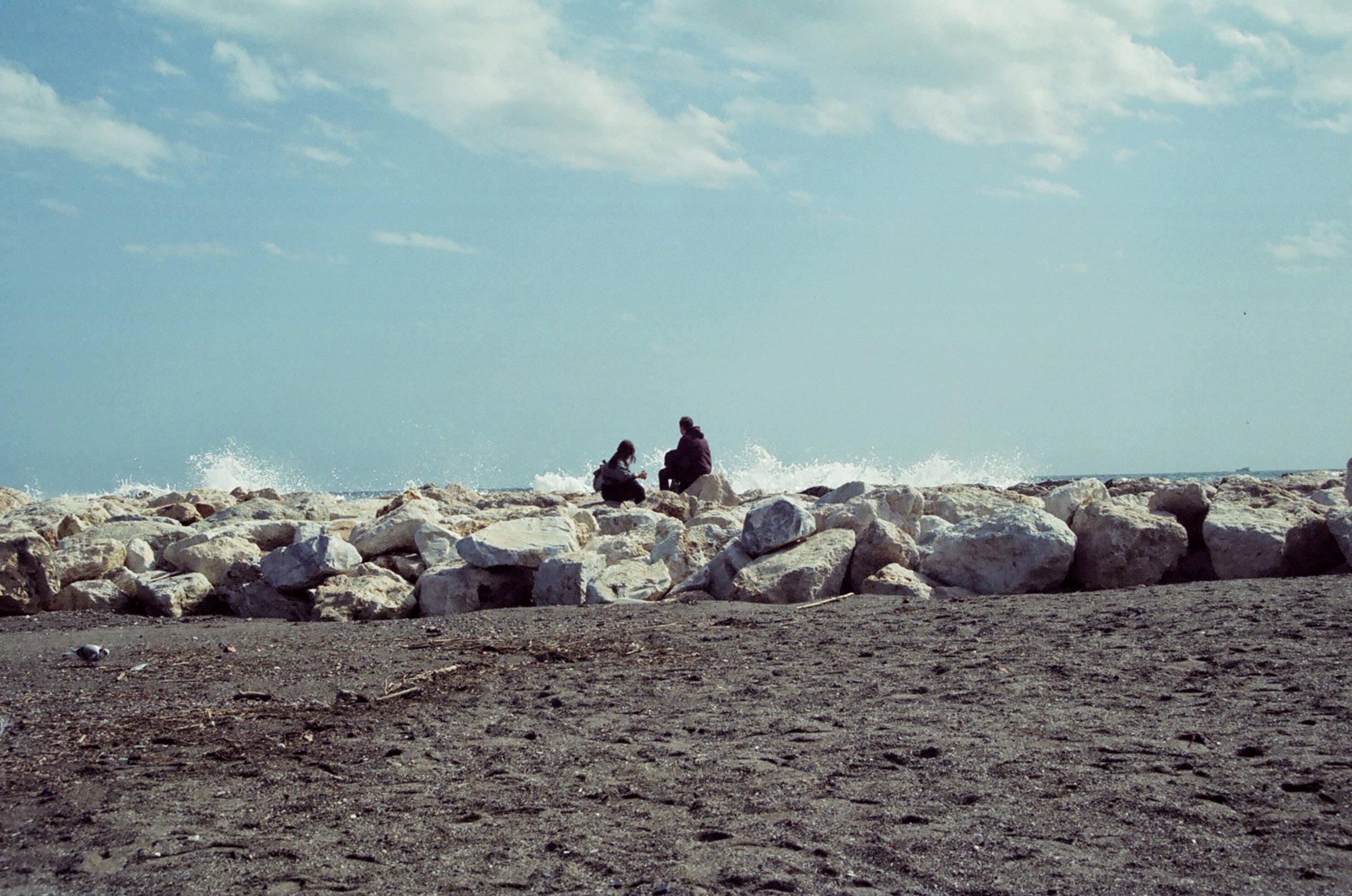 Two people sitting on rocks by the ocean, with waves crashing in the background and a cloudy sky overhead.