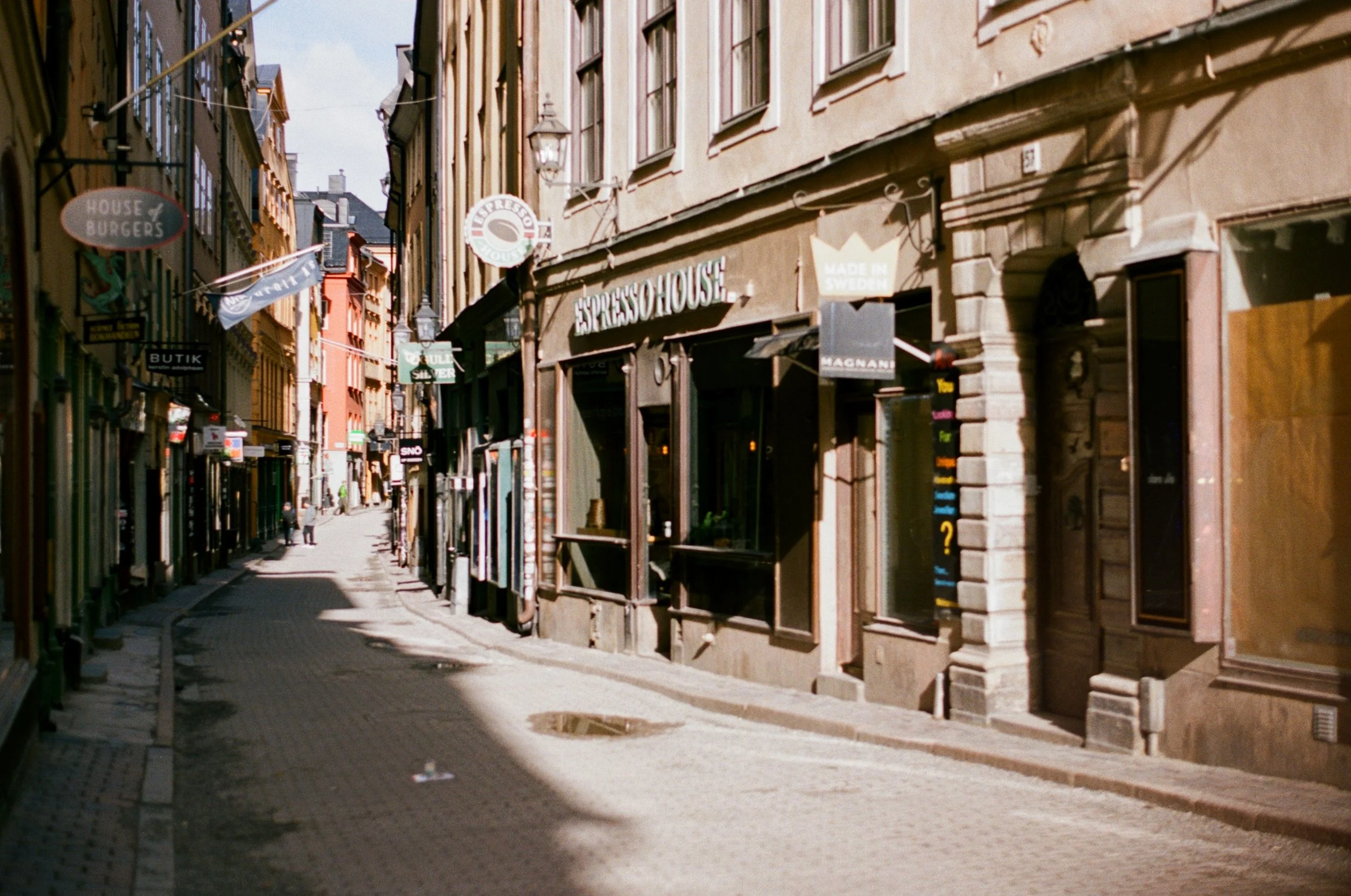 A narrow cobblestone street in a European city with tall buildings on both sides, featuring signs for various shops and cafes, including 'Espresso House' and 'House of Burgers', with a few pedestrians in the distance.