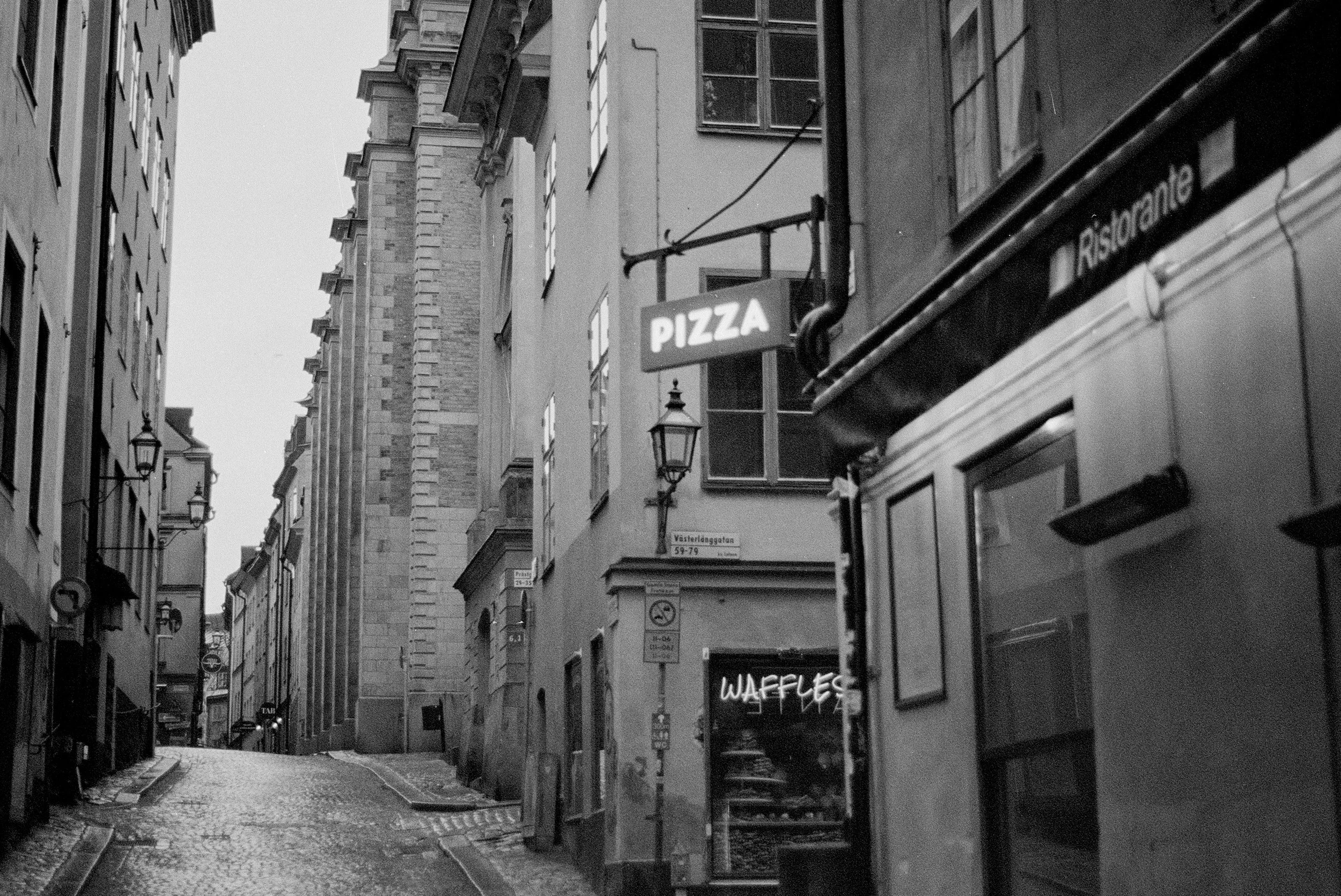 A black and white photo of a narrow cobblestone street lined with tall buildings. The street has a sign for pizza and a neon sign for waffles. There are lamp posts along the buildings, and the street appears to be slightly wet.