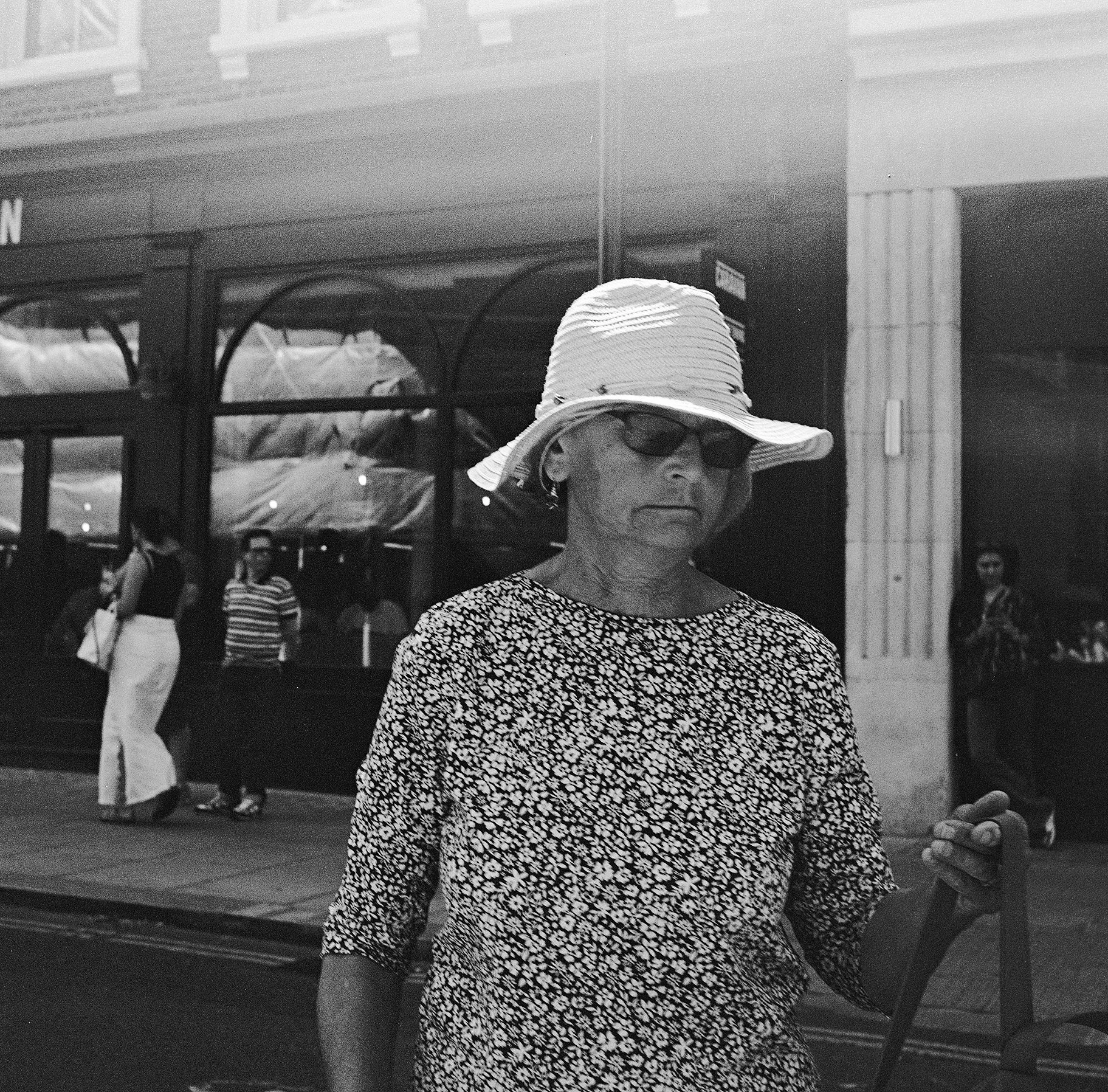 A woman wearing a wide-brimmed hat and sunglasses walks on a city sidewalk, with a storefront and other pedestrians in the background.