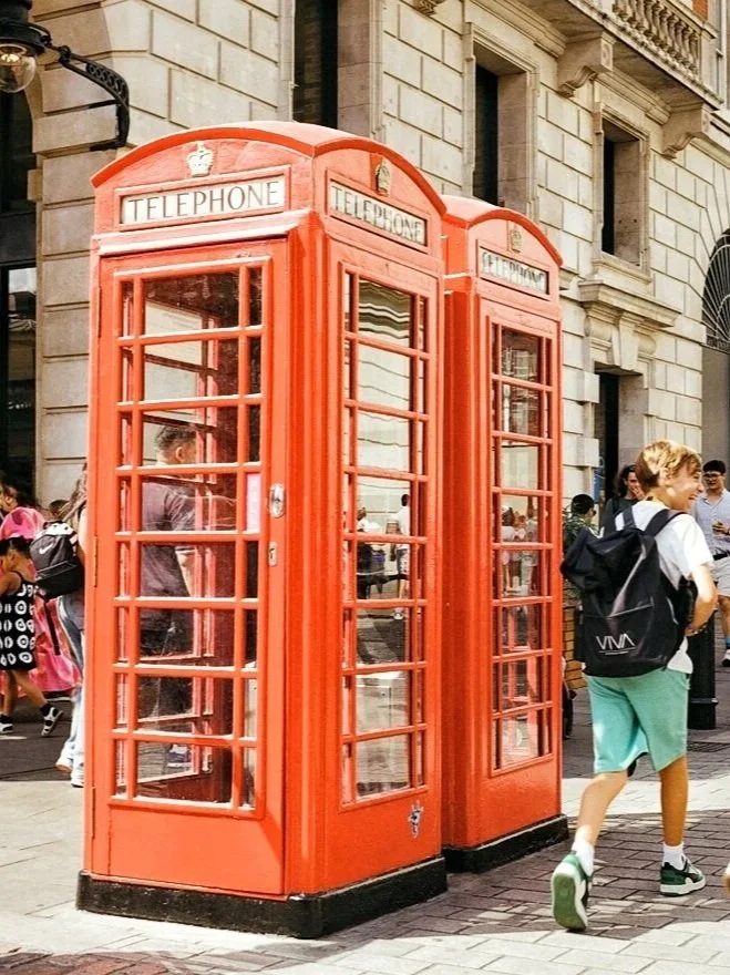 Two red British-style telephone booths on a busy city street with people walking by.