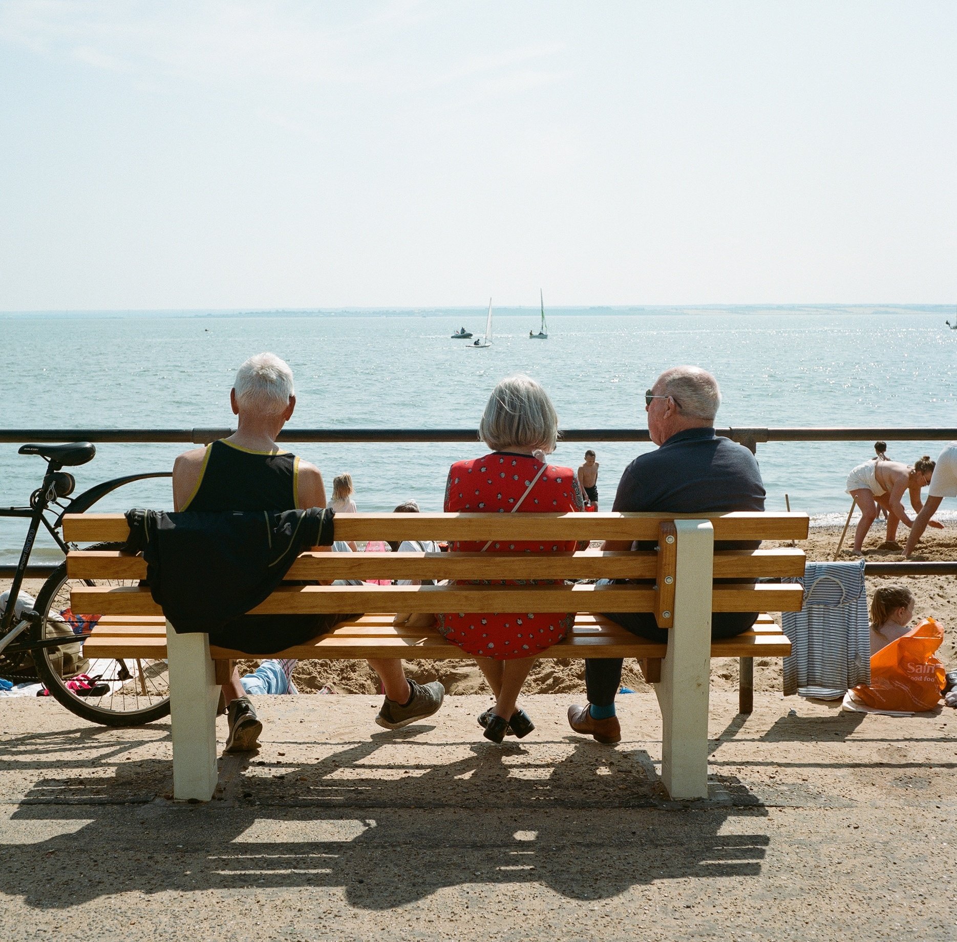 Three elderly people sitting on a wooden bench facing the water at a beach, with sailboats on the horizon and people on the sand.