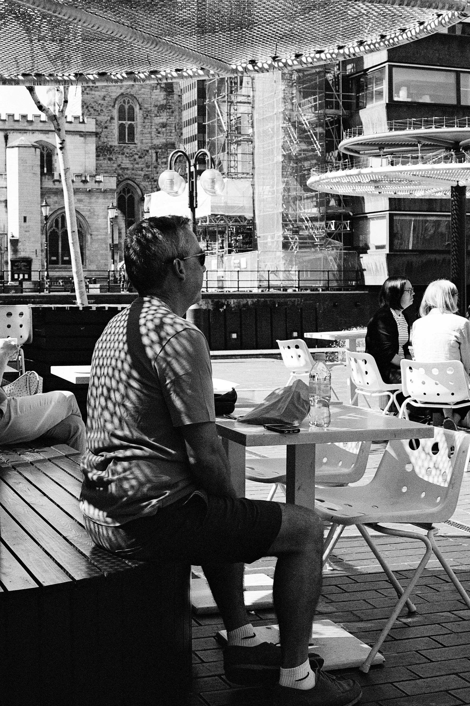 A man sitting at an outdoor table in a cafe, with a cityscape and construction in the background, black-and-white photo.