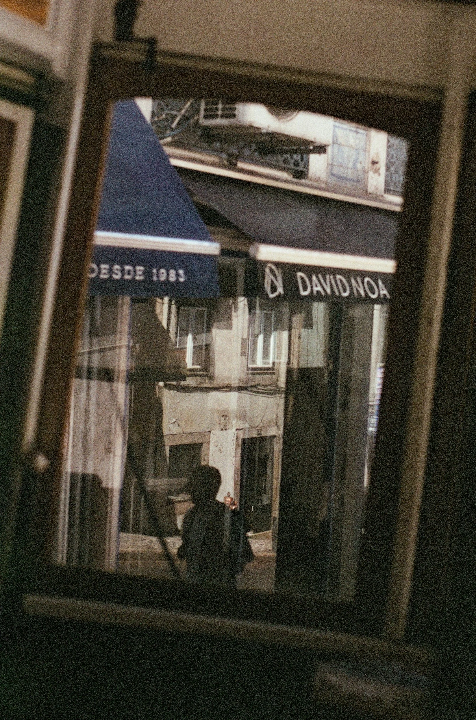 View of a person walking outside through a window, with signs for "DAVID NOA" and "DESDE 1935," a building in the background, and a street scene.