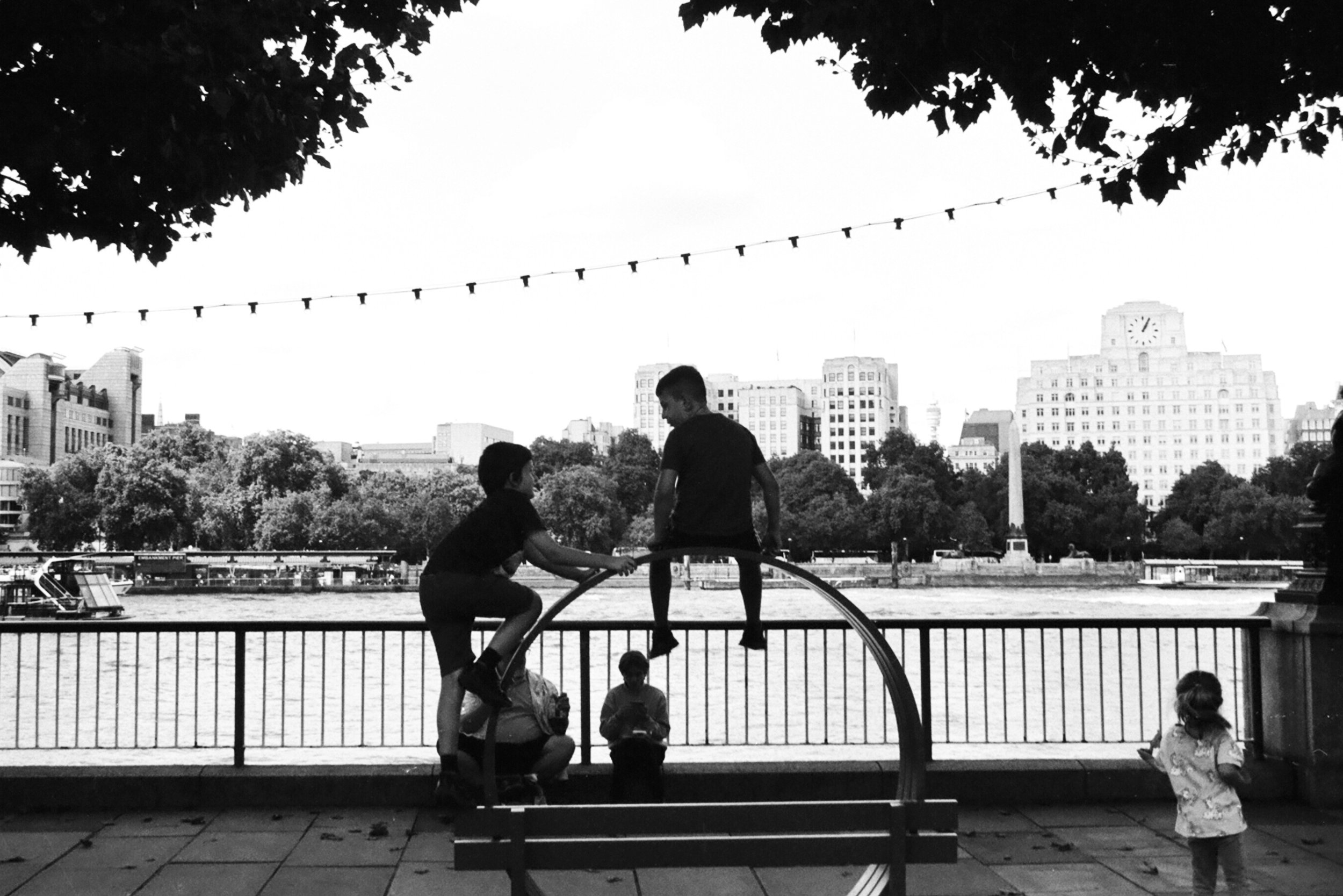 Children playing on a park structure with a river and city skyline in the background, some people sitting near a railing, trees, and a clock tower in the distance.