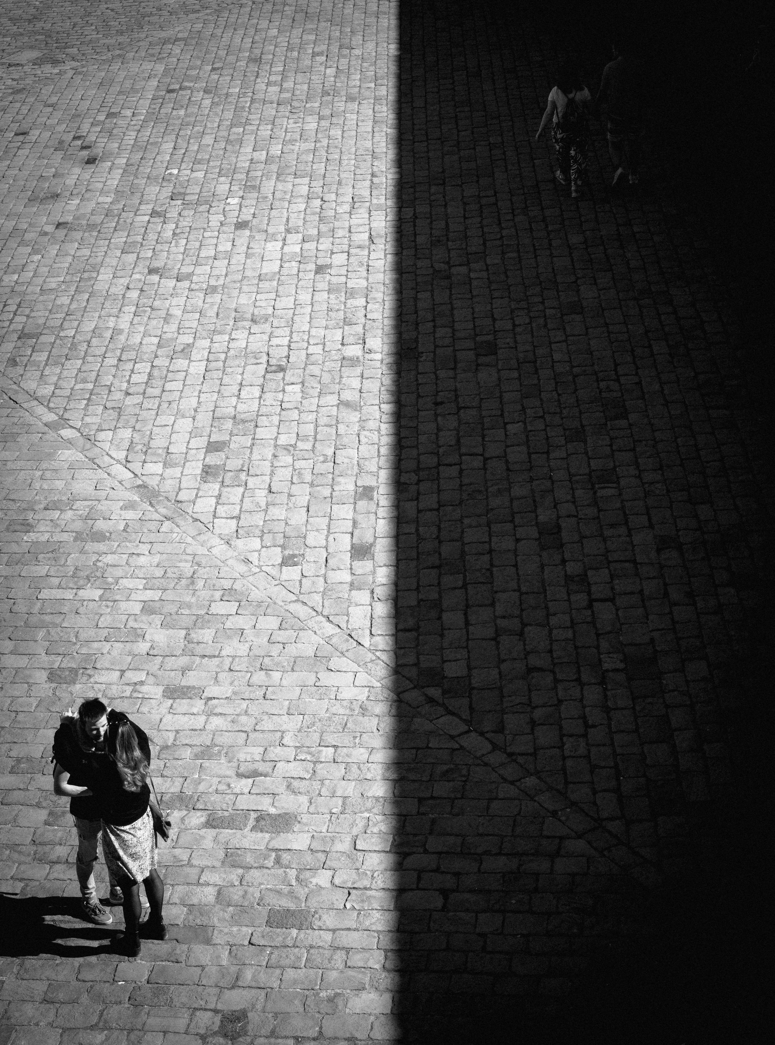 Two couples are standing on cobblestone pavement, separated by a sharp shadow dividing the lighter and darker areas of the ground.
