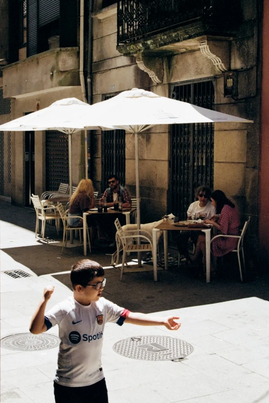 People sitting at outdoor tables under umbrellas in a street café during the day, with a child playing in the foreground.