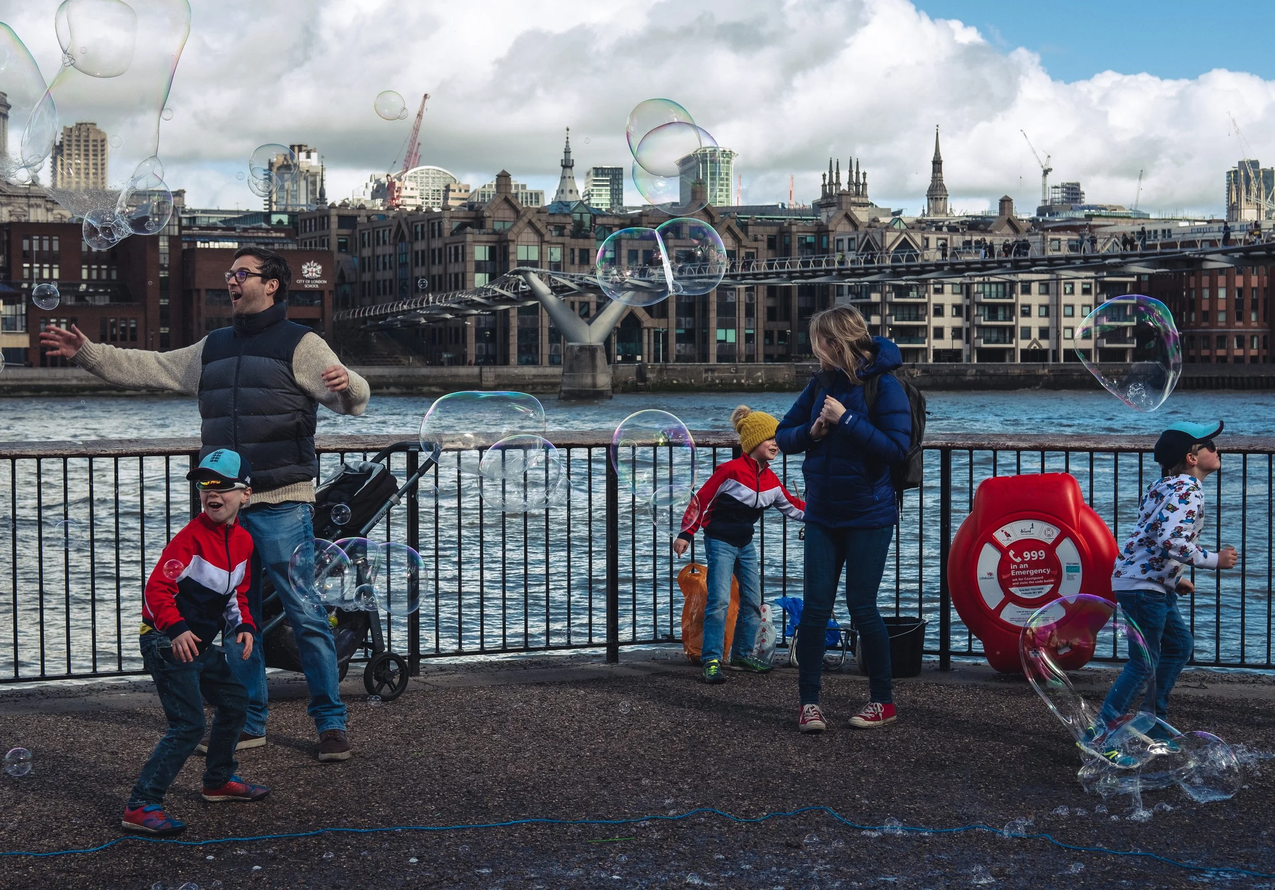 People playing with bubbles along a riverfront with city buildings, a bridge, and cloudy sky in the background.