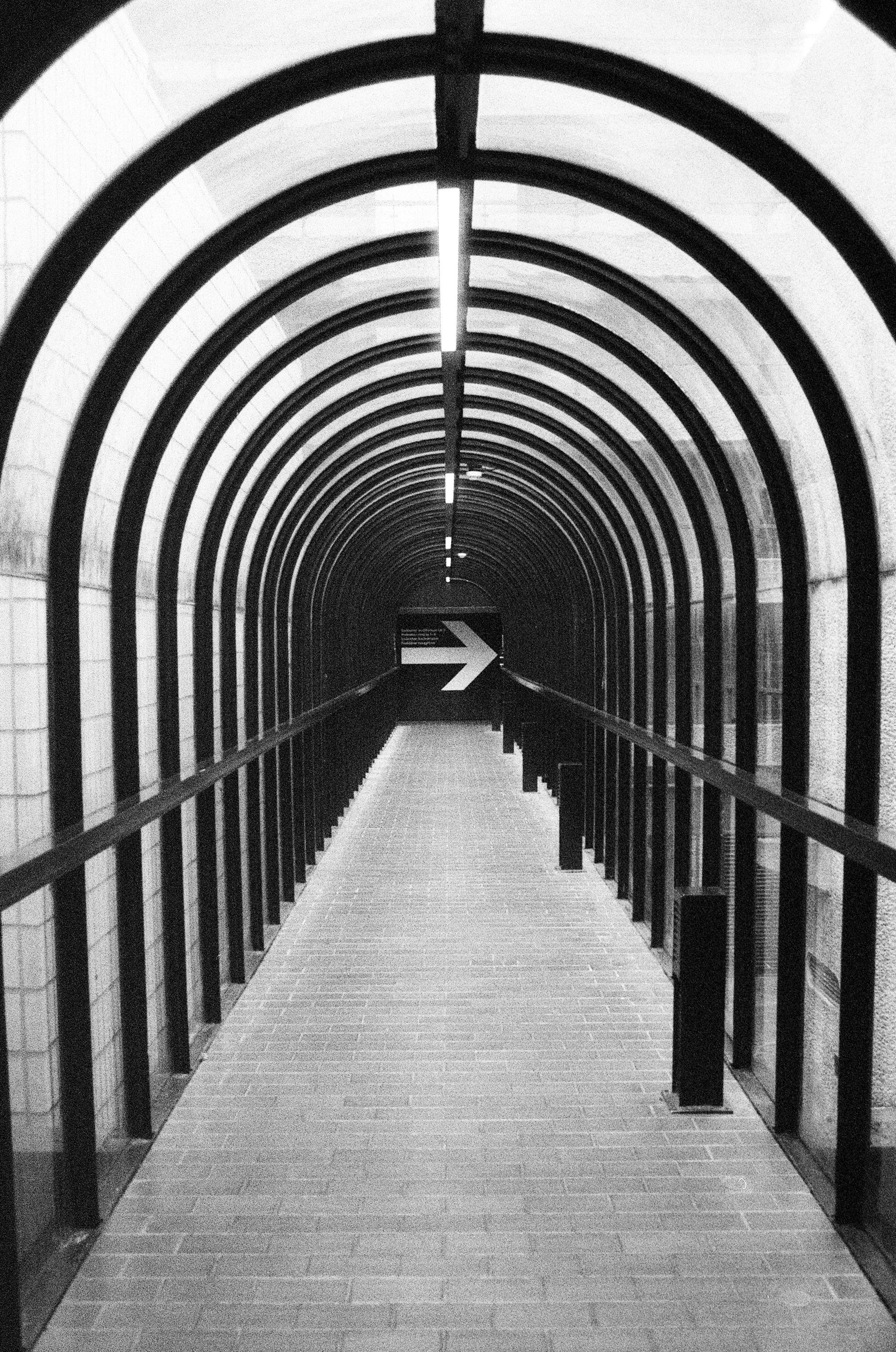 A black and white photo of a tunnel with a walkway, metal arches overhead, and an arrow pointing right.