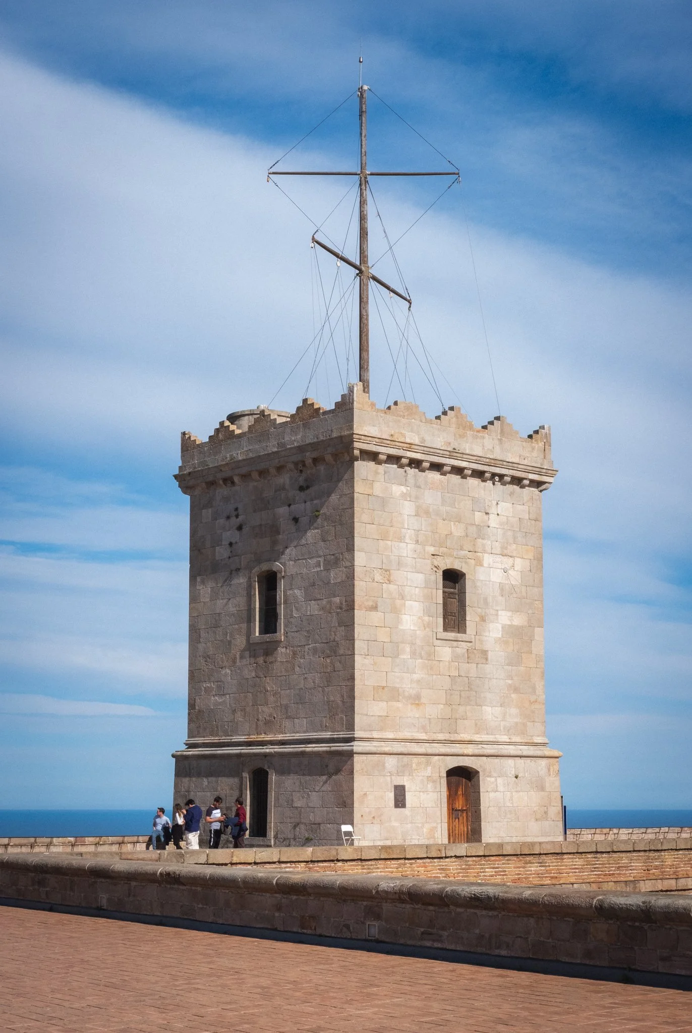 A tall stone tower with three small windows and a wooden door, topped with a mast and rigging, overlooking the ocean on a clear day.
