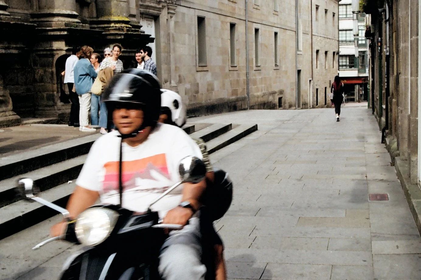 A person riding a scooter wearing a helmet on a city street with a group of people standing near a building on the left and a woman walking away on the right.