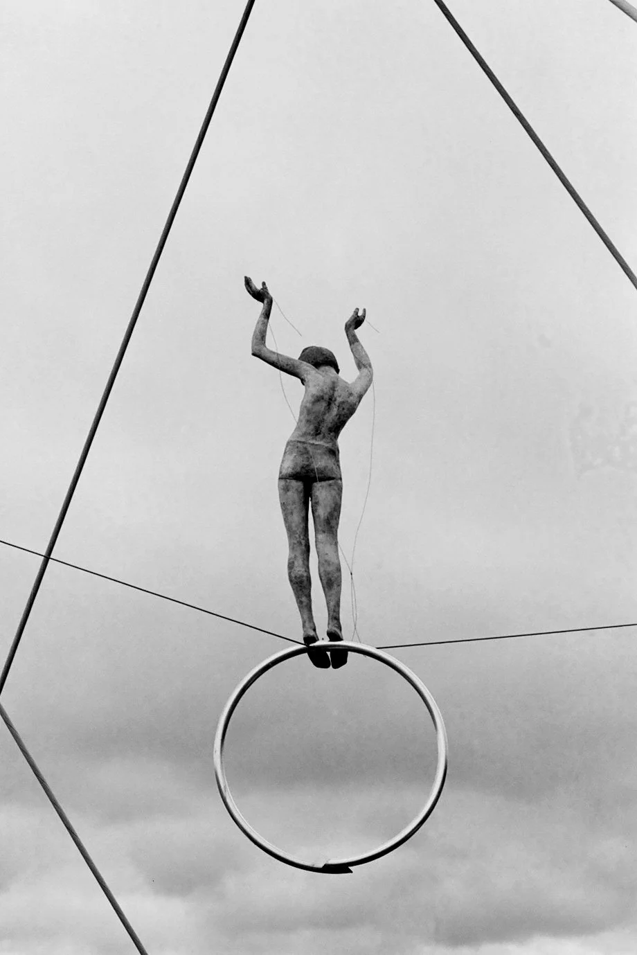 A trapeze artist in a costume, with arms raised, hanging upside down and balancing on a circular hoop, with a cloudy sky in the background.