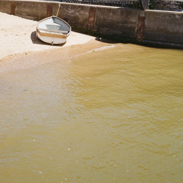 A small white boat resting on a sandy shore next to murky yellowish water, with a large rusted metal structure in the background.