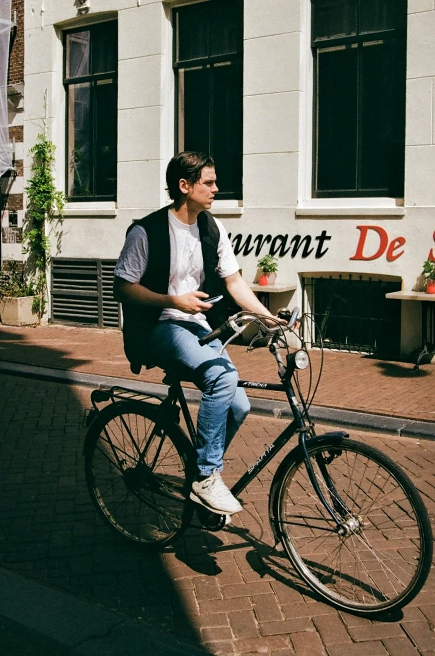 A young man riding a black bicycle on a city street in front of a building with large windows and part of a sign that reads "Restaurant." He is wearing a gray T-shirt, black vest, and jeans, and is looking to his left while holding a smartphone.