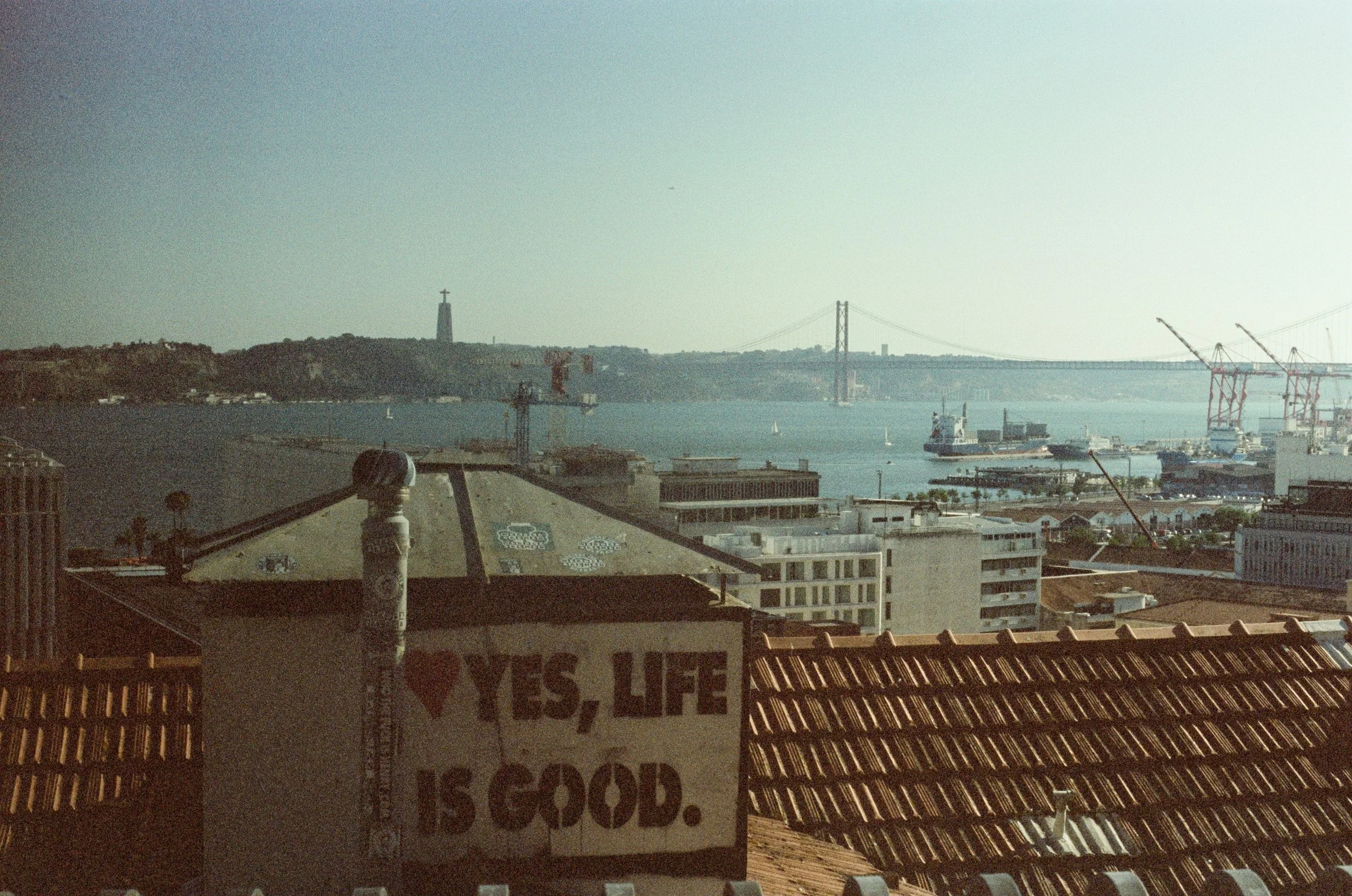 Cityscape with a bay, cranes, a bridge, and the statue of Christ the Redeemer in the distance, with a rooftop in the foreground featuring a mural that reads 'I love YES, LIFE IS GOOD'.