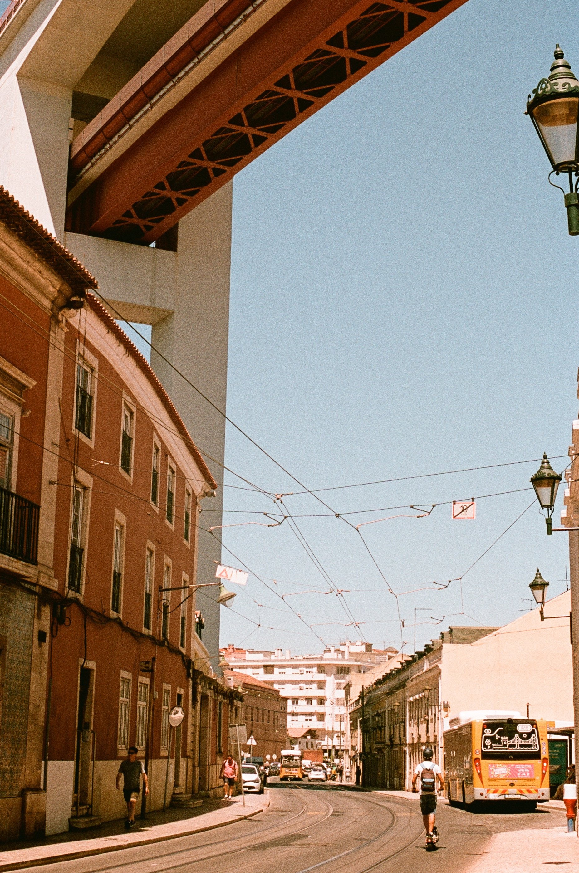 A city street scene with a tram, pedestrians, and a bridge overhead, under a clear blue sky.