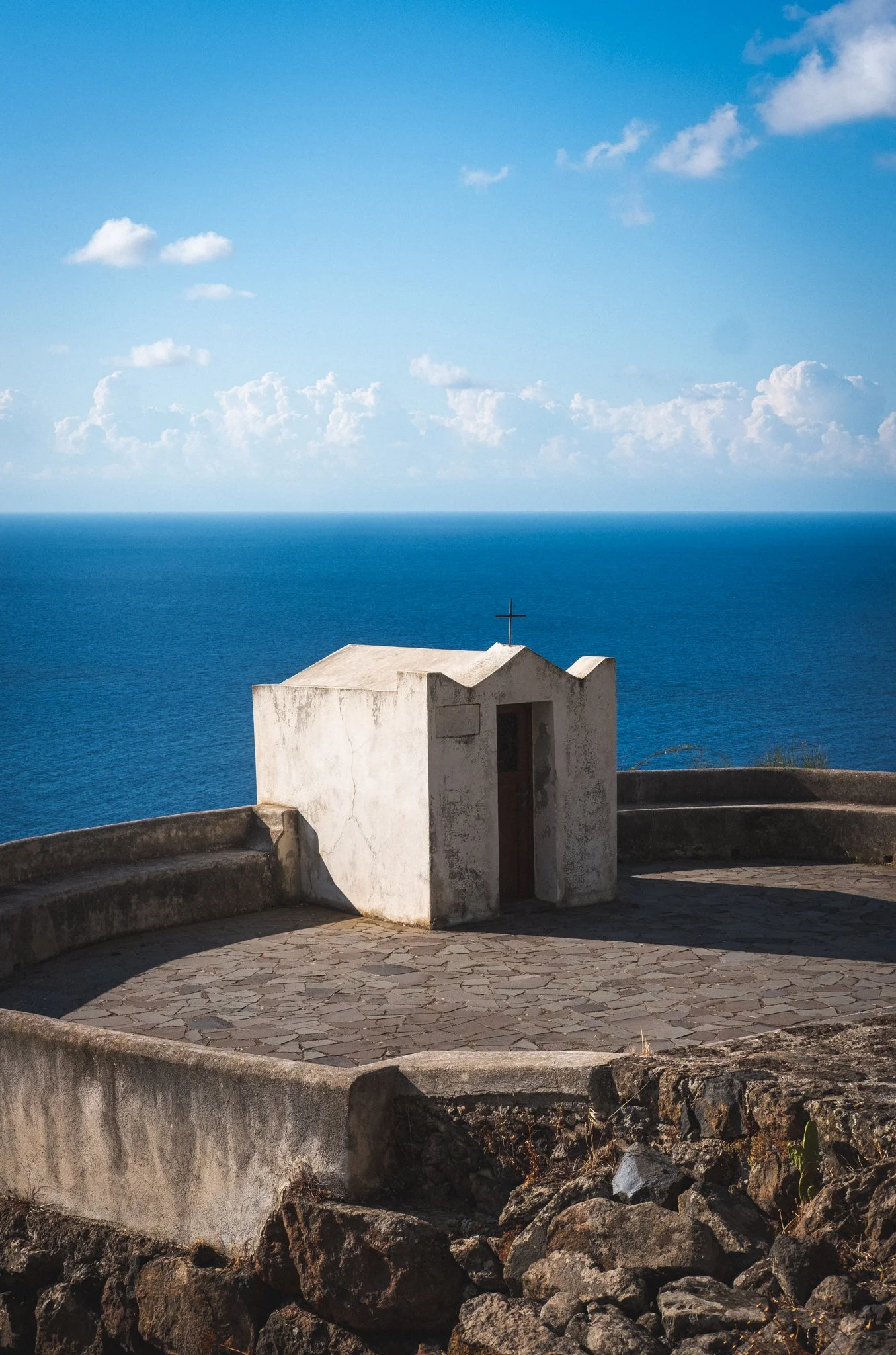Small white church with a cross on top, overlooking the ocean under a blue sky with clouds.