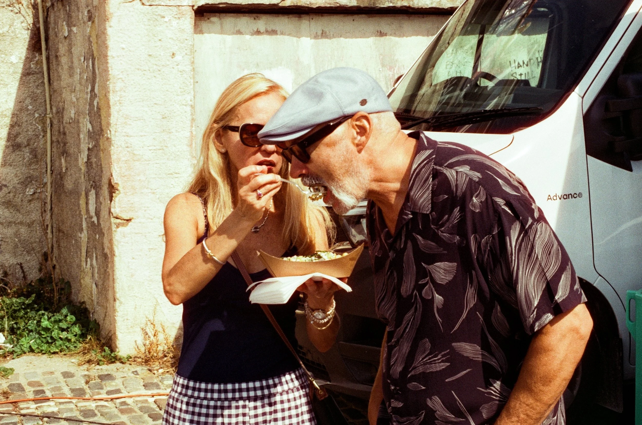 A woman feeding an older man with a fork outdoors near a white van and a stone wall.