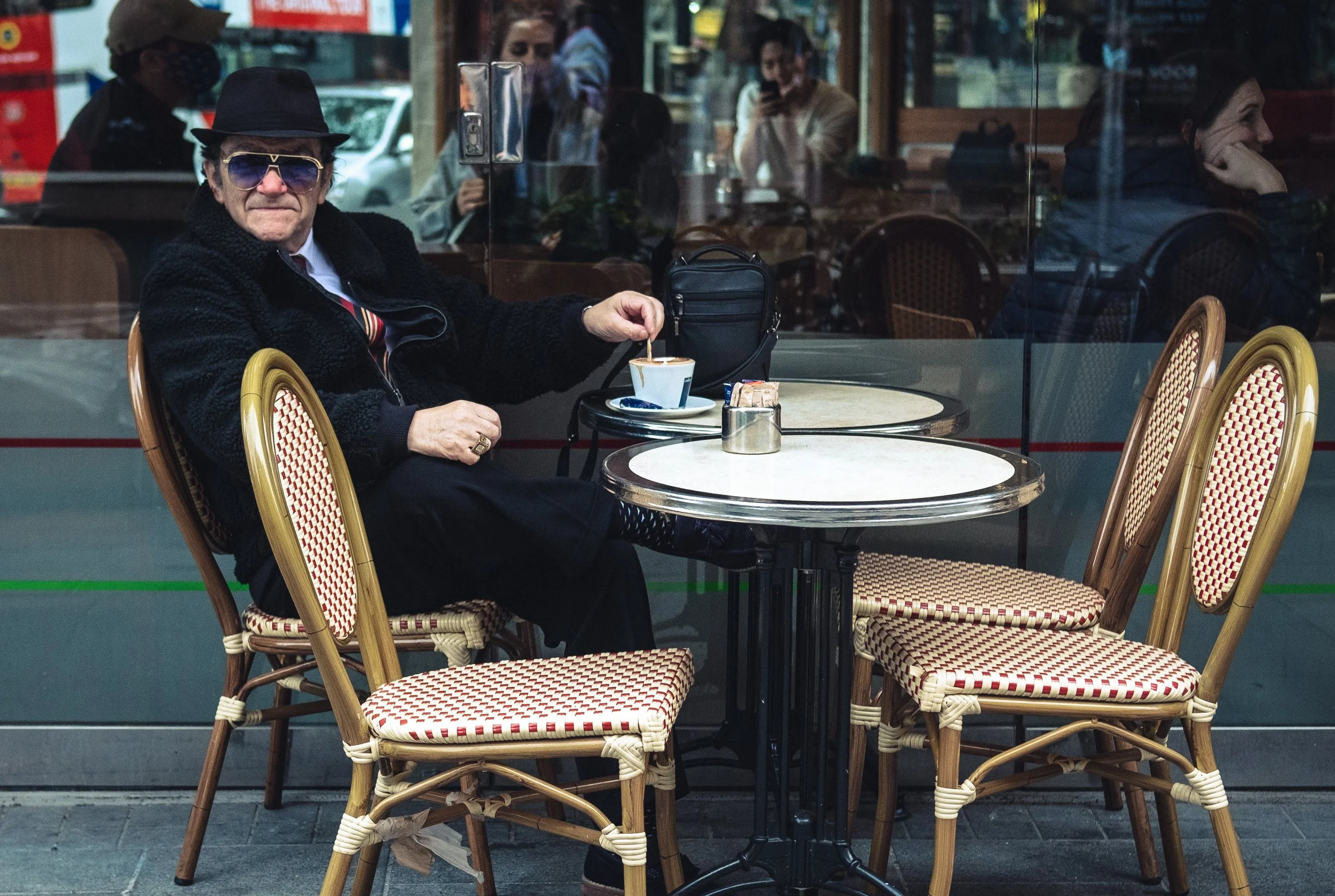 A man in a black hat, sunglasses, and a black jacket sitting outside a cafe at a table, stirring a cup of coffee, with several empty chairs around him and other people reflected in the window behind.