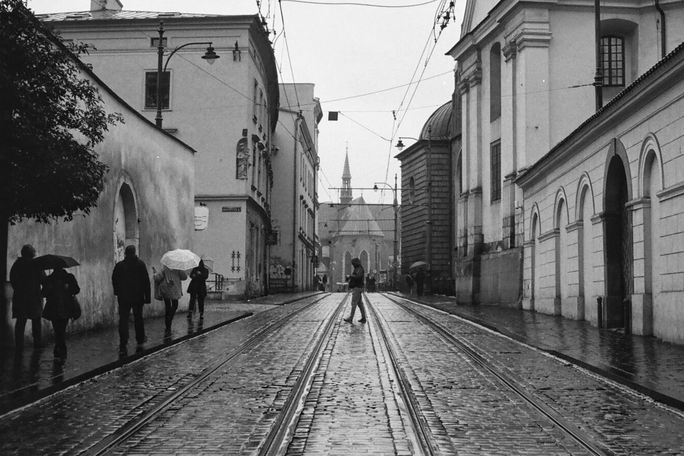 Black and white photo of a wet city street with tram tracks, pedestrians with umbrellas, and historic buildings on both sides. A church steeple is visible in the background.